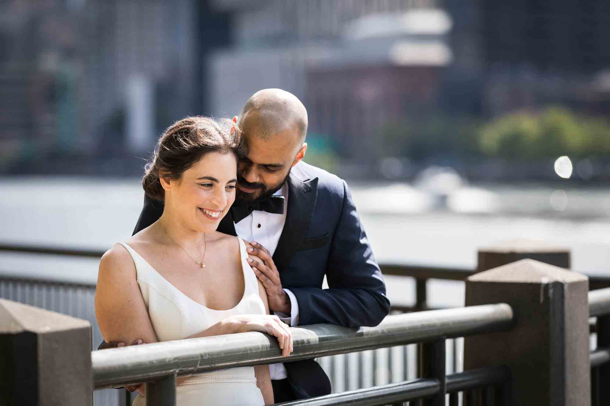 Bride and groom cuddling along railing on waterfront in front of NYC skyline on Roosevelt Island