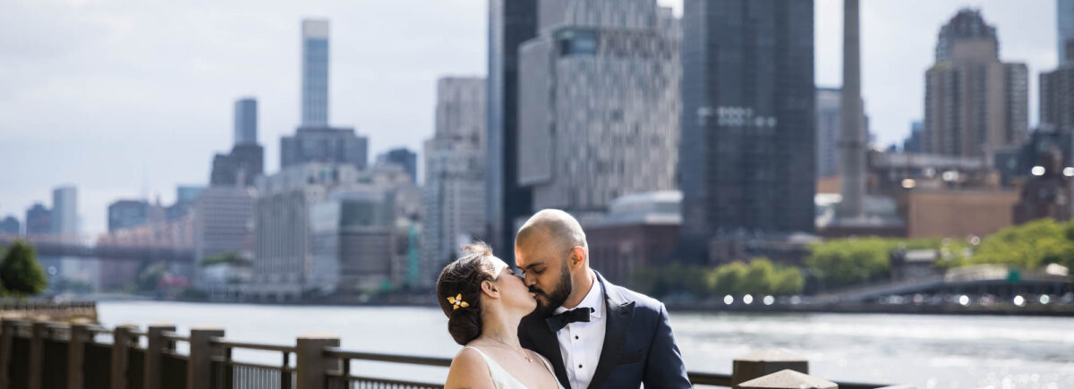 Bride and groom kissing on railing along waterfront in front of NYC skyline on Roosevelt Island