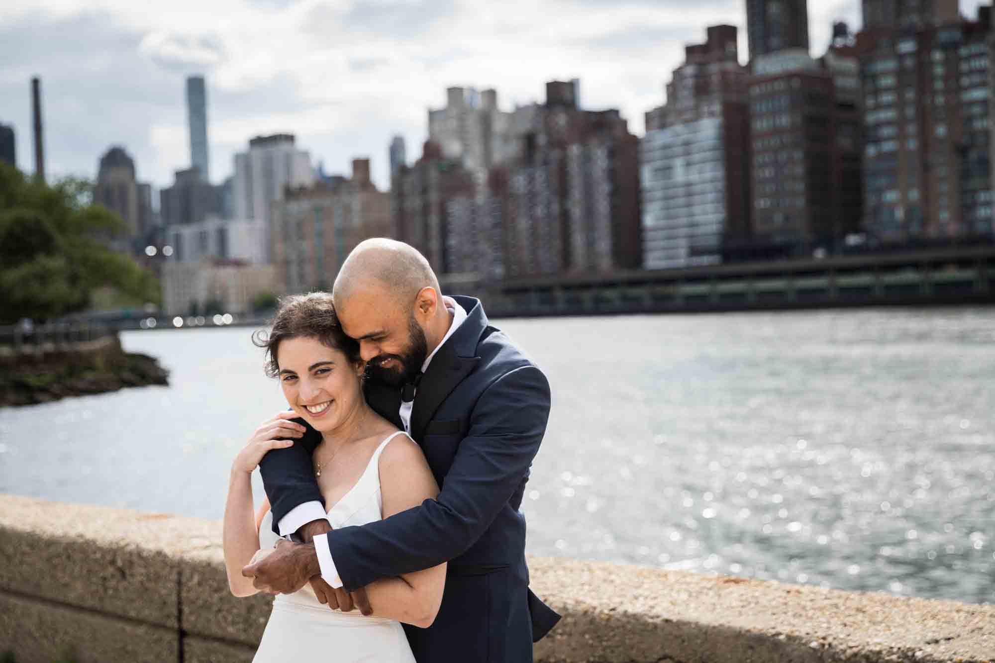 Groom hugging bride from behind in front of NYC skyline on Roosevelt Island