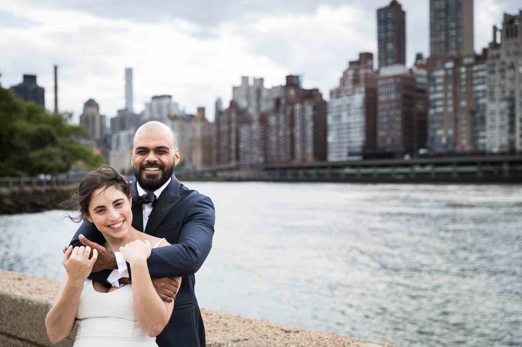 Groom hugging bride from behind in front of NYC skyline on Roosevelt Island