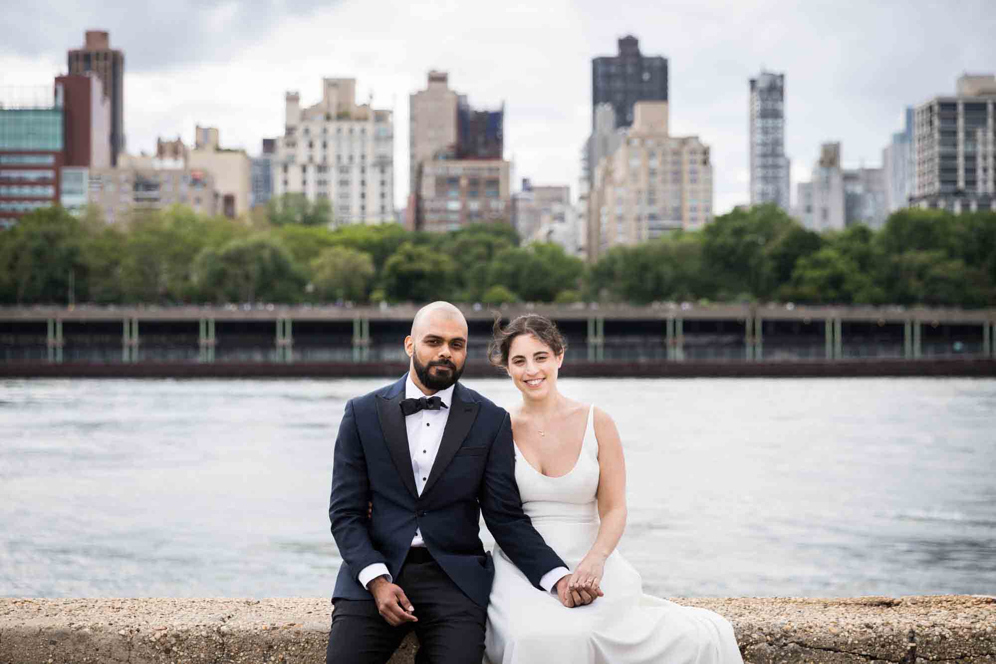 Bride and groom holding hands and sitting on stone wall in front of NYC skyline on Roosevelt Island