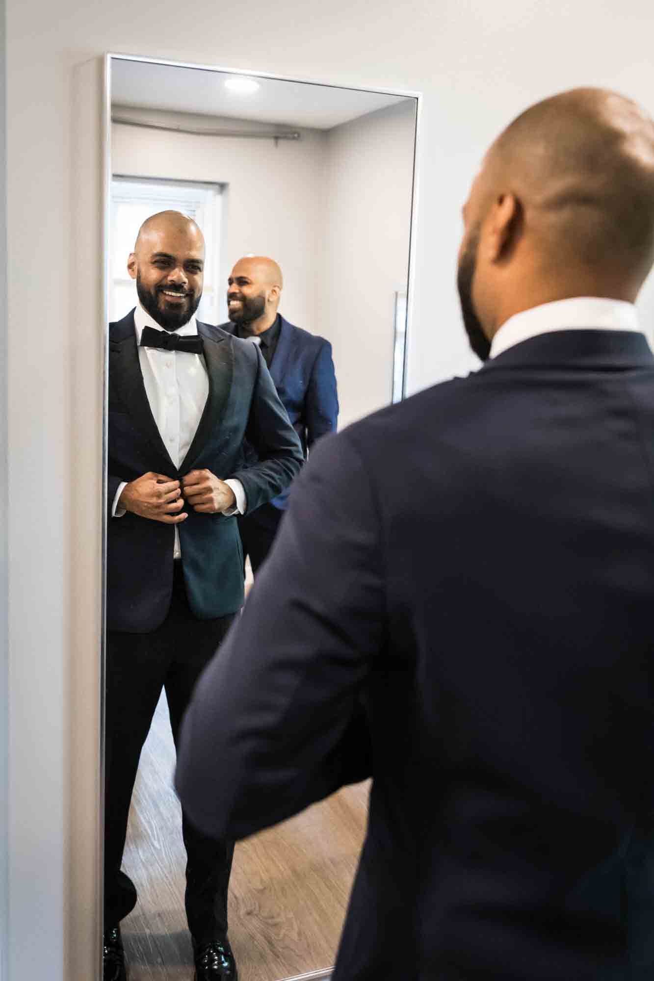 Groom adjusting tux jacket in mirror with man in background at a Sanctuary Roosevelt Island wedding