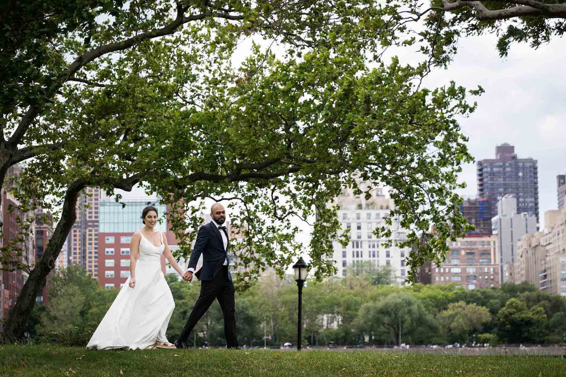 Bride and groom holding hands and walking in park in front of NYC skyline on Roosevelt Island