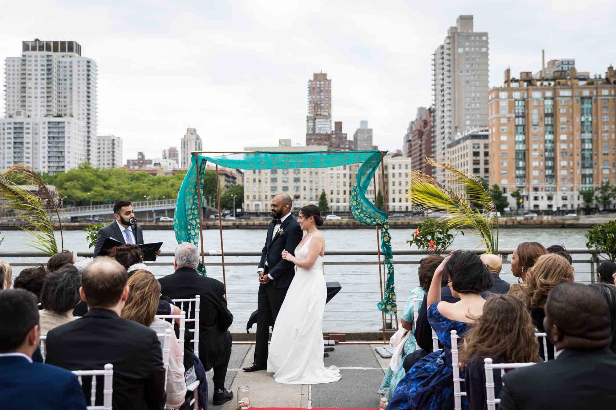 Bride and groom listening to officiant under green chuppah at a Sanctuary Roosevelt Island wedding