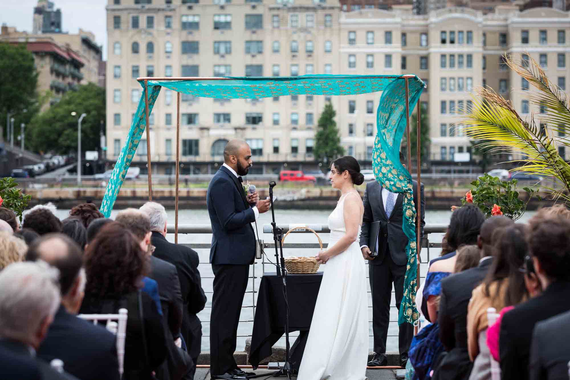 Bride and groom saying vows under green chuppah in front of NYC skyline at a Sanctuary Roosevelt Island wedding