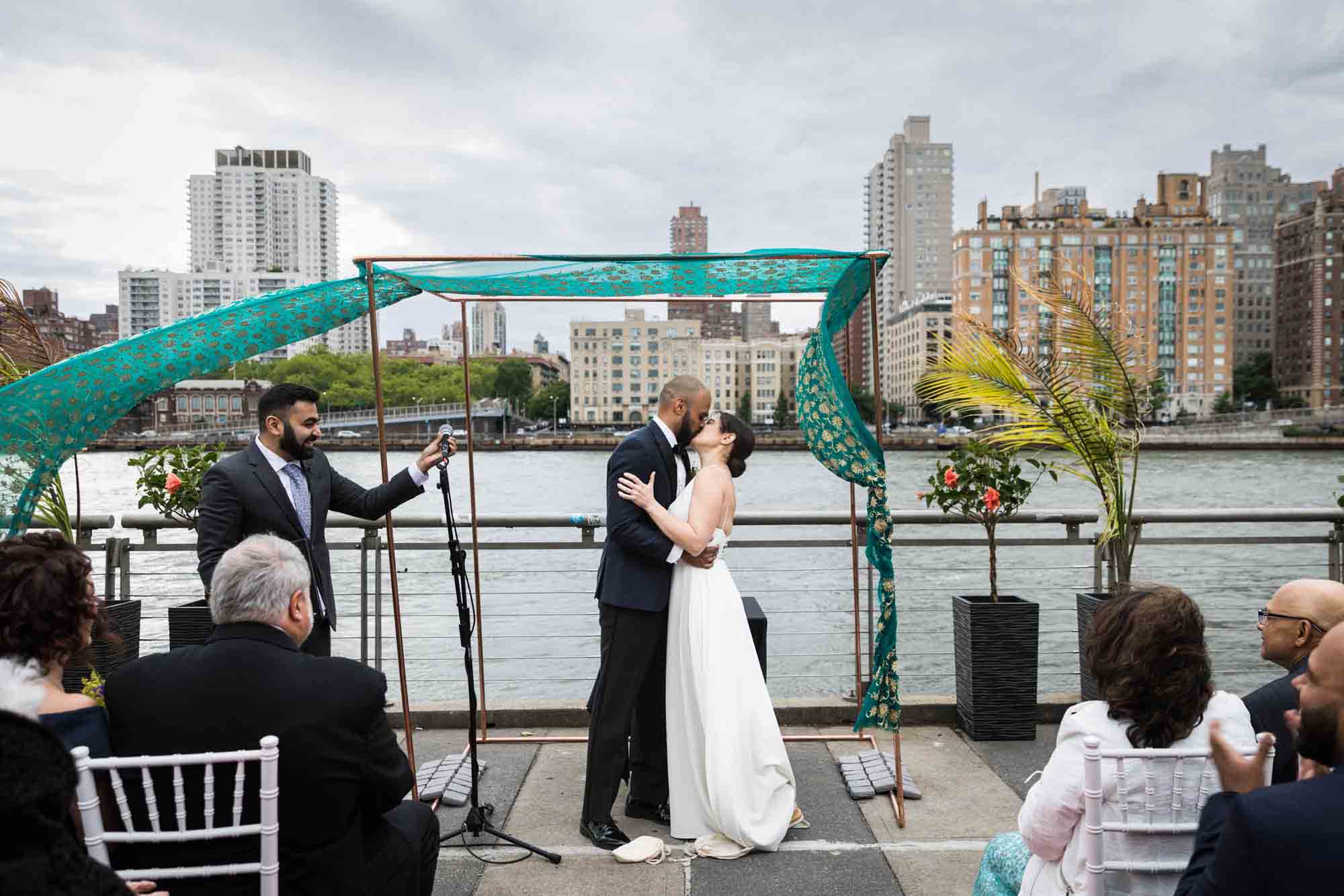 Bride and groom kissing under green chuppah during outdoors ceremony at a Sanctuary Roosevelt Island wedding