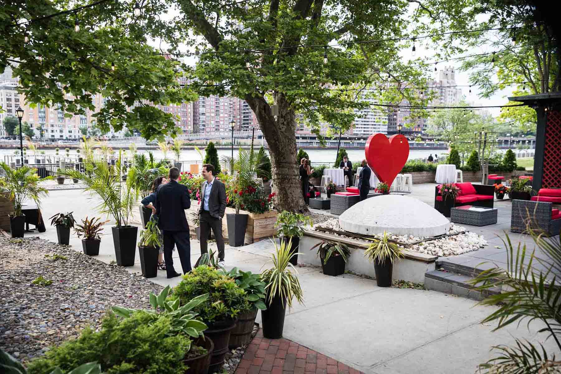 Guests talking on outdoor patio with red heart sculpture and plants at a Sanctuary Roosevelt Island wedding