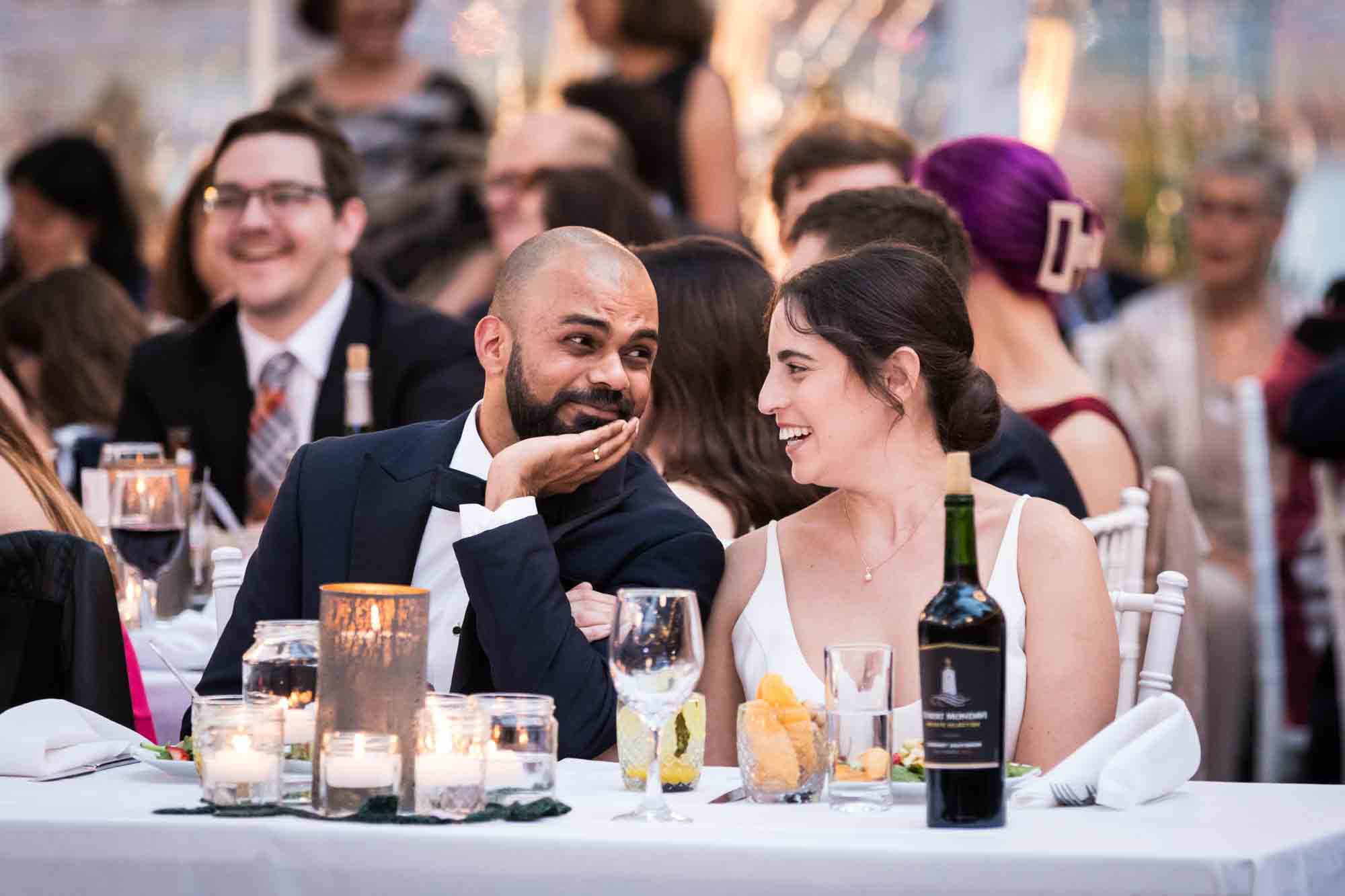 Bride and groom looking at each other sitting at table behind wine bottle at a Sanctuary Roosevelt Island wedding