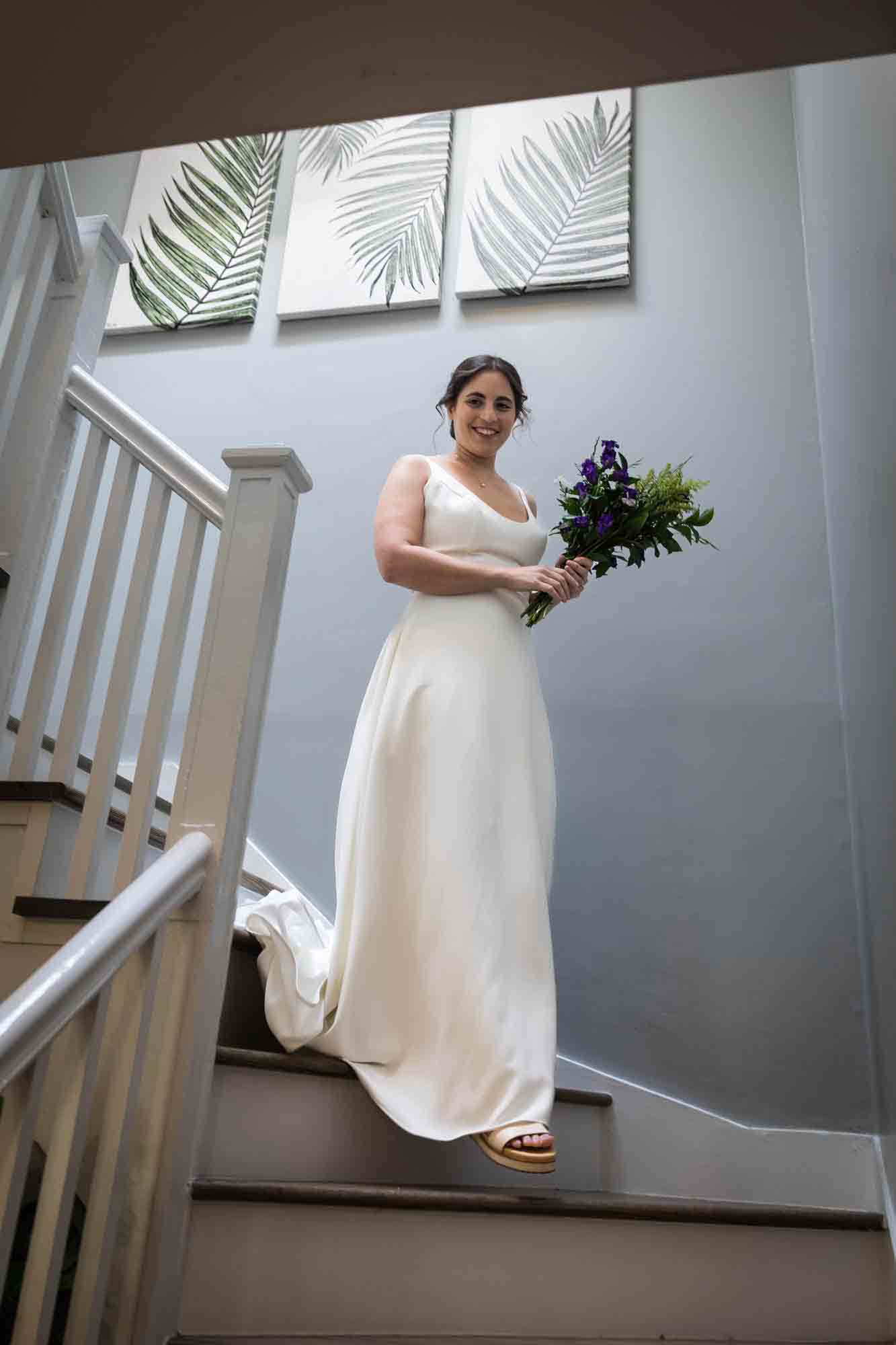 Bride wearing white sleeveless wedding dress holding bouquet of flowers walking down stairs at a Sanctuary Roosevelt Island wedding