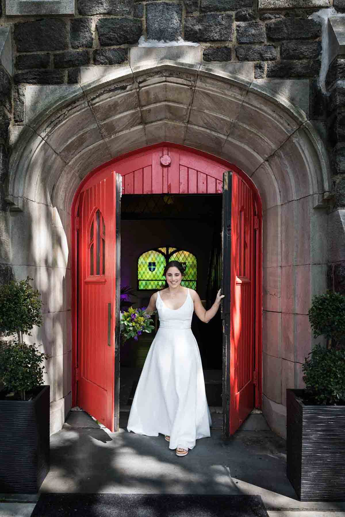 Bride wearing white dress and holding flower bouquet walking out of red doors at a Sanctuary Roosevelt Island wedding