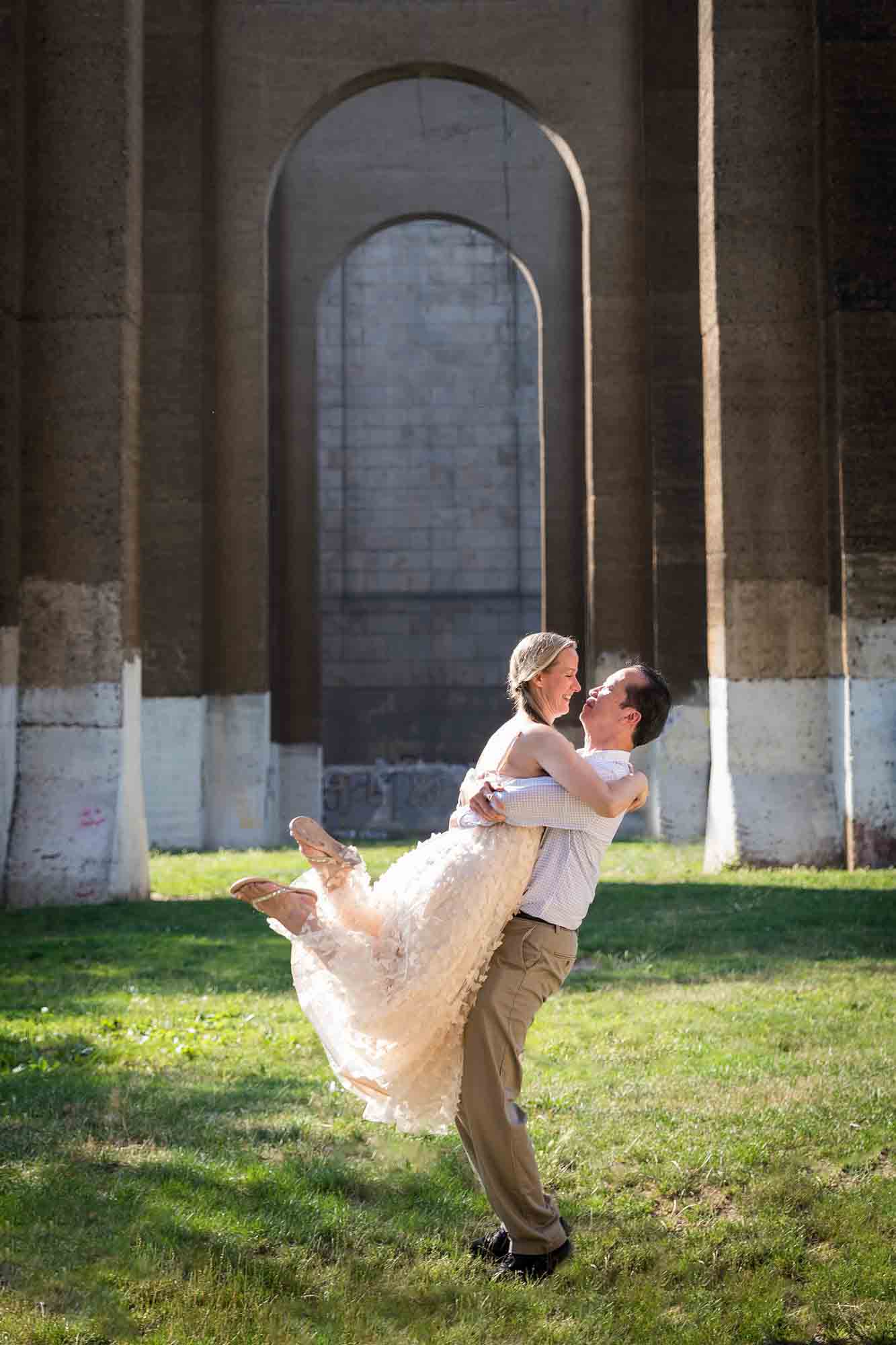 Astoria Park engagement photos of a man lifting woman up under the Hell Gate Bridge