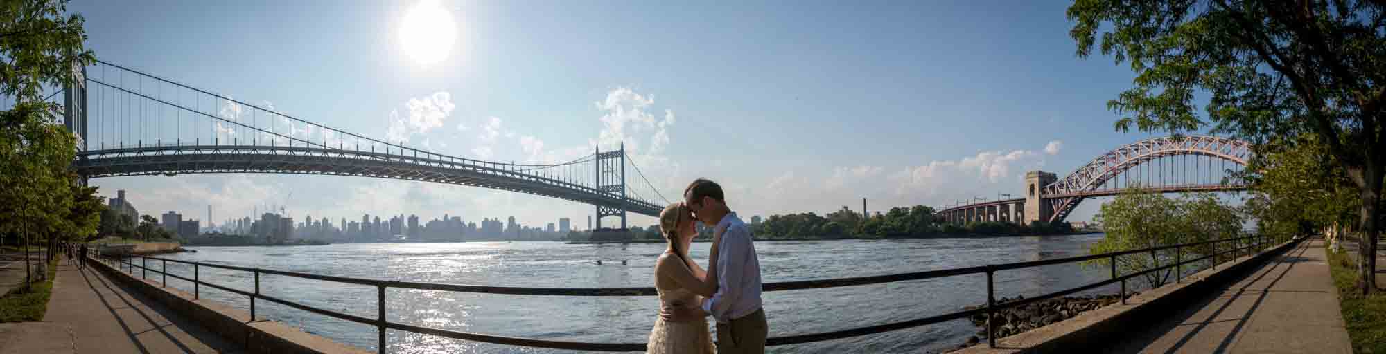 Panoramic photo of couple hugging by waterfront in front of Queensborough Bridge and Hell Gate Bridge