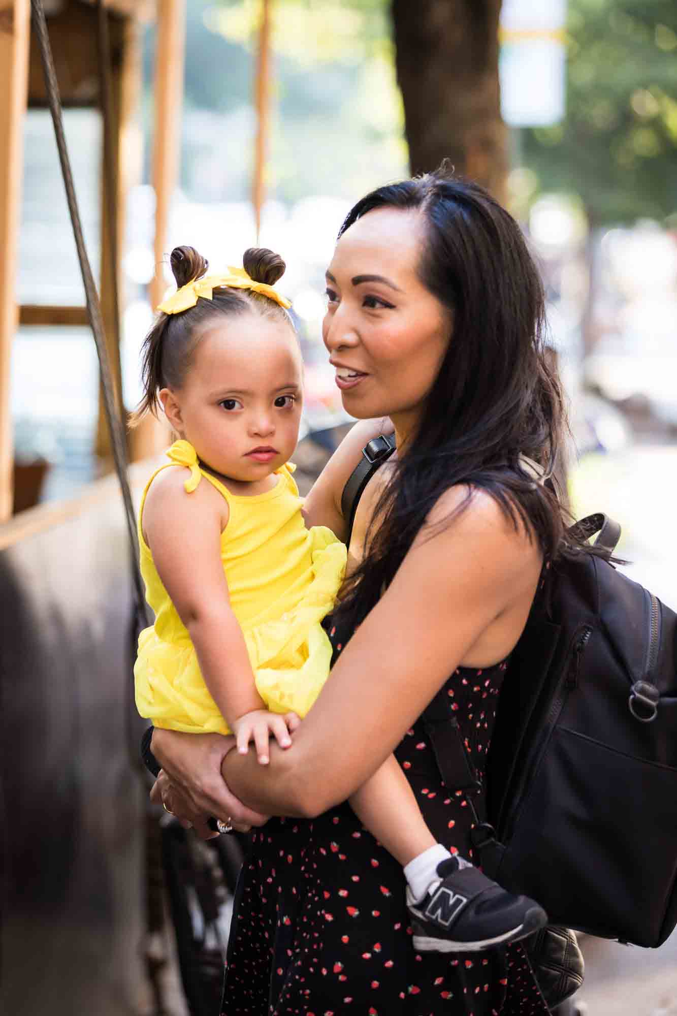 Mother holding daughter wearing yellow dress during rehearsal dinner at an outdoor restaurant