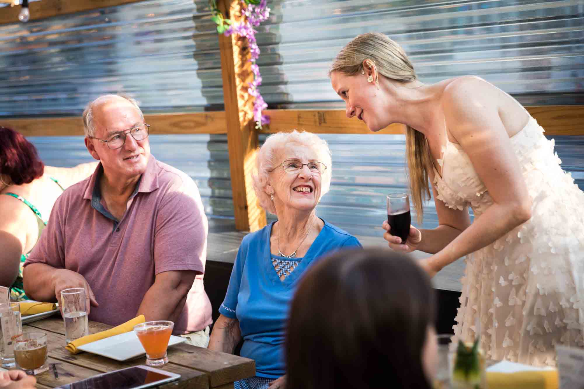 Bride speaking to seated grandmother and father during rehearsal dinner at an outdoor restaurant