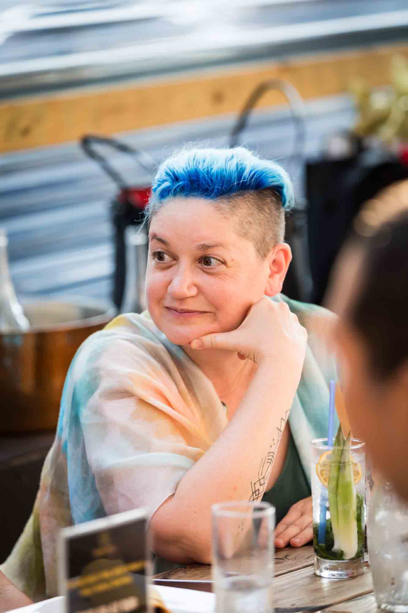 Woman with blue hair and hand to chin during rehearsal dinner at an outdoor restaurant