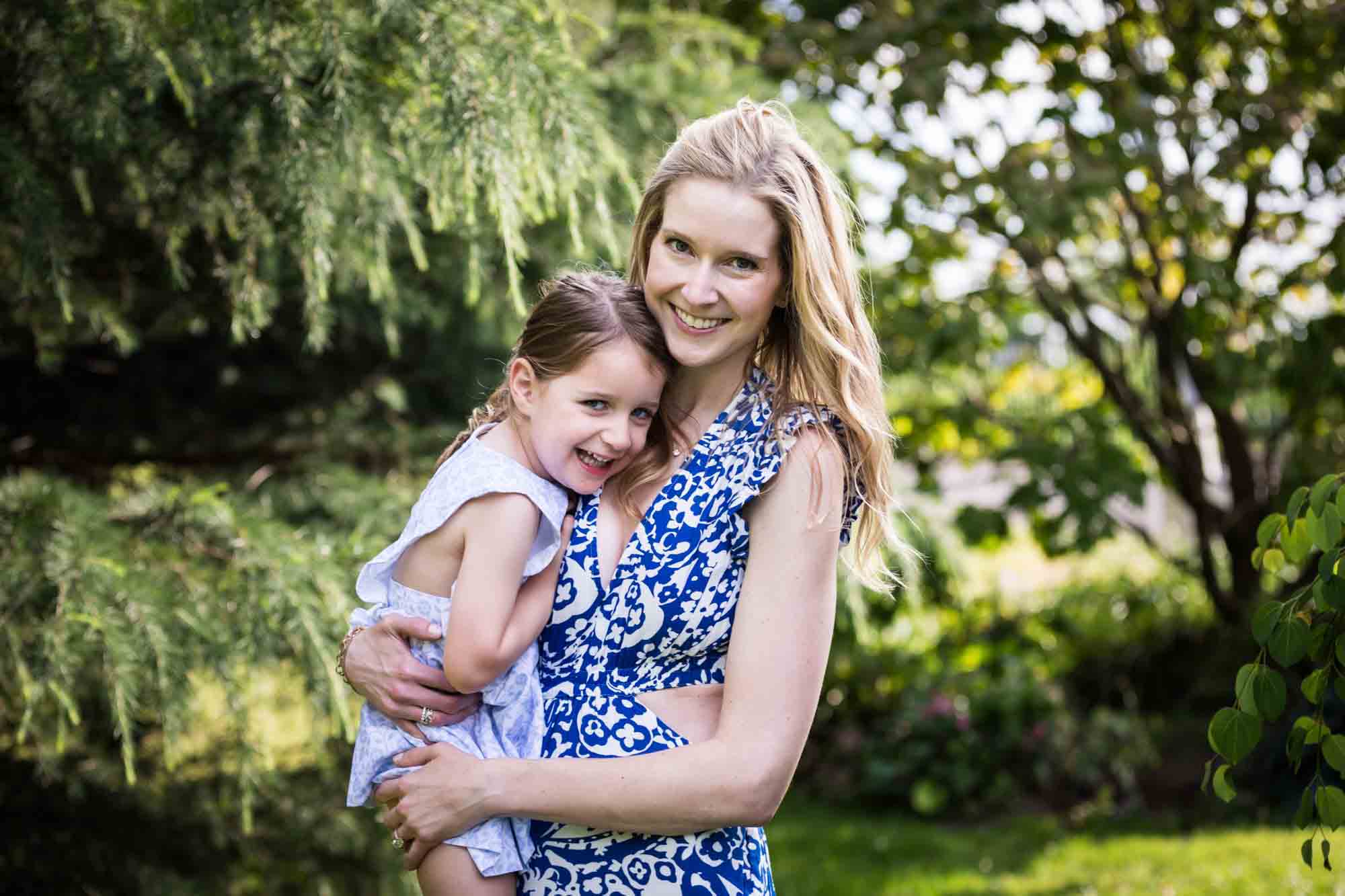 Blonde mother in blue dress holding little girl in front of trees during a Narrows Botanical Gardens family portrait session