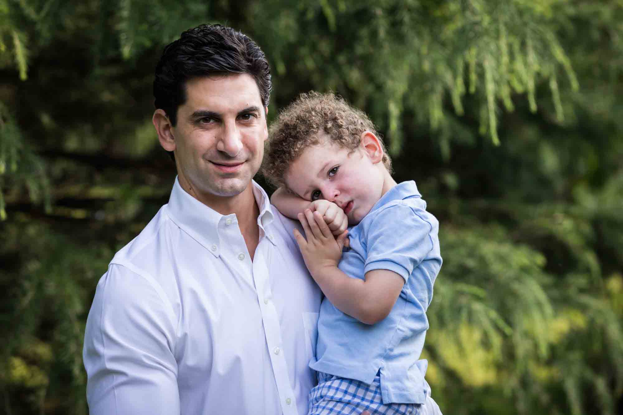 Man with brown hair and white shirt holding little boy with curly hair in front of a tree during a Narrows Botanical Gardens family portrait session