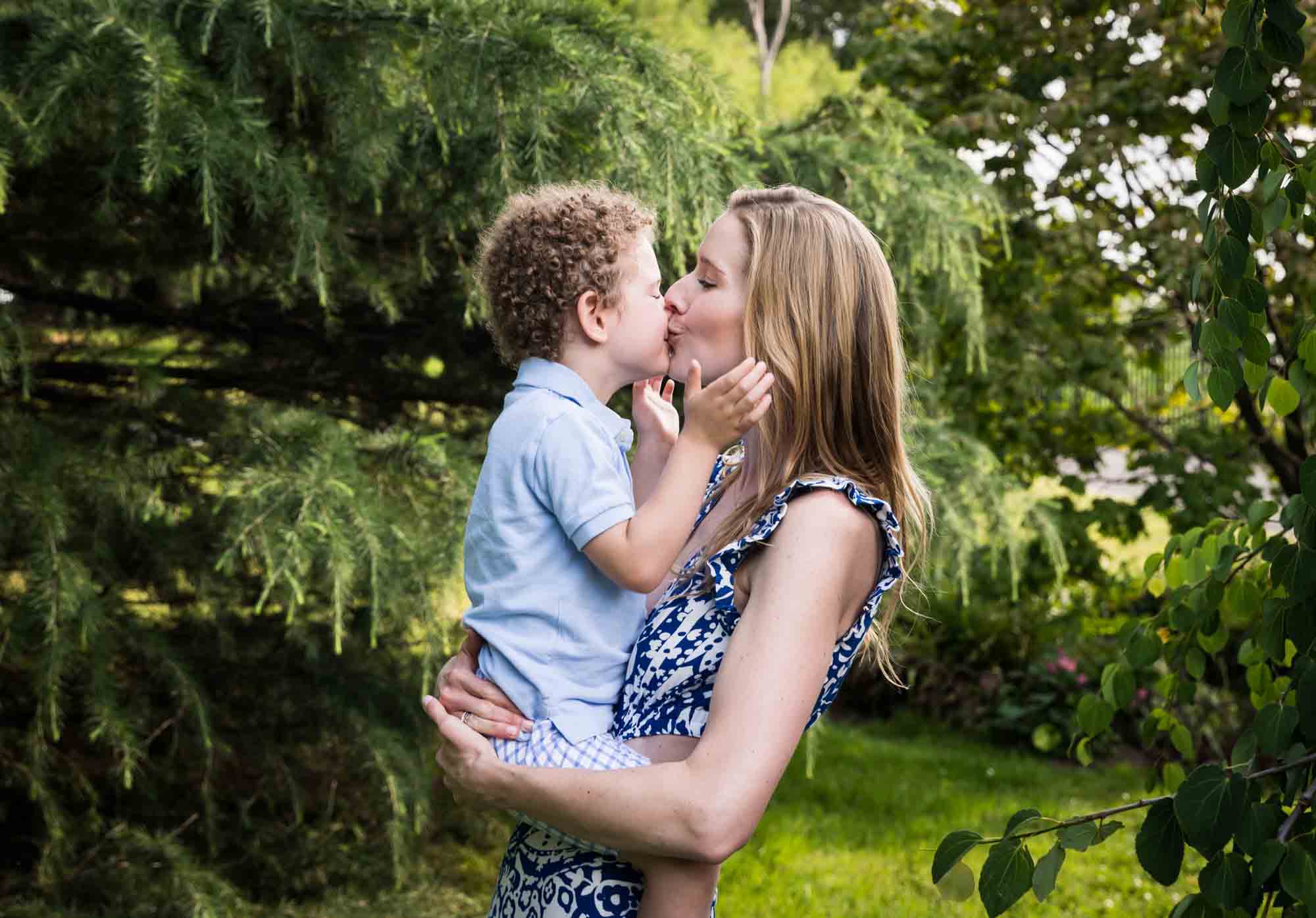 Blonde mother kissing little boy with curly hair in front of a tree during a Narrows Botanical Gardens family portrait session