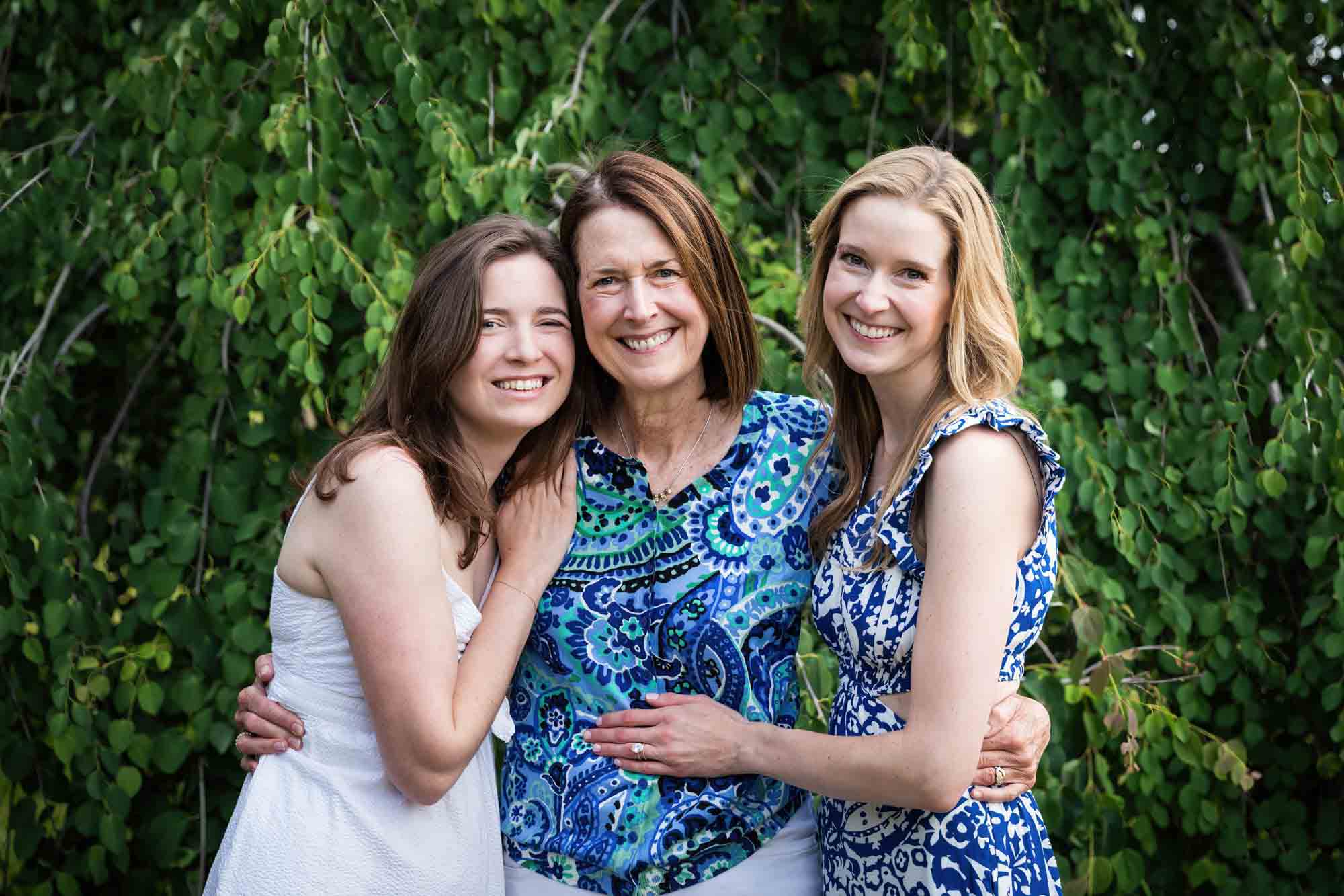 An older mother and two grown daughters hugging in front of a tree during a Narrows Botanical Gardens family portrait session