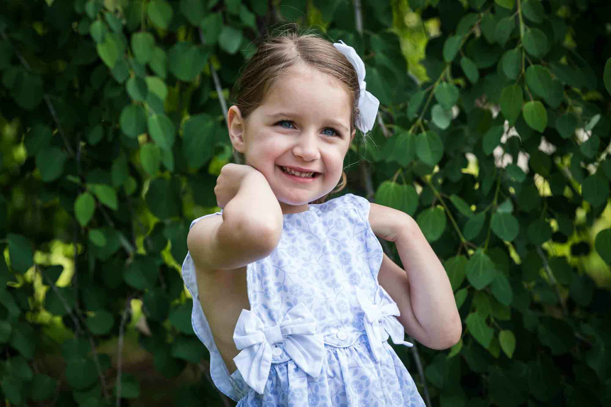 Smiling little girl wearing blue dress standing in front of tree during a Narrows Botanical Gardens family portrait session