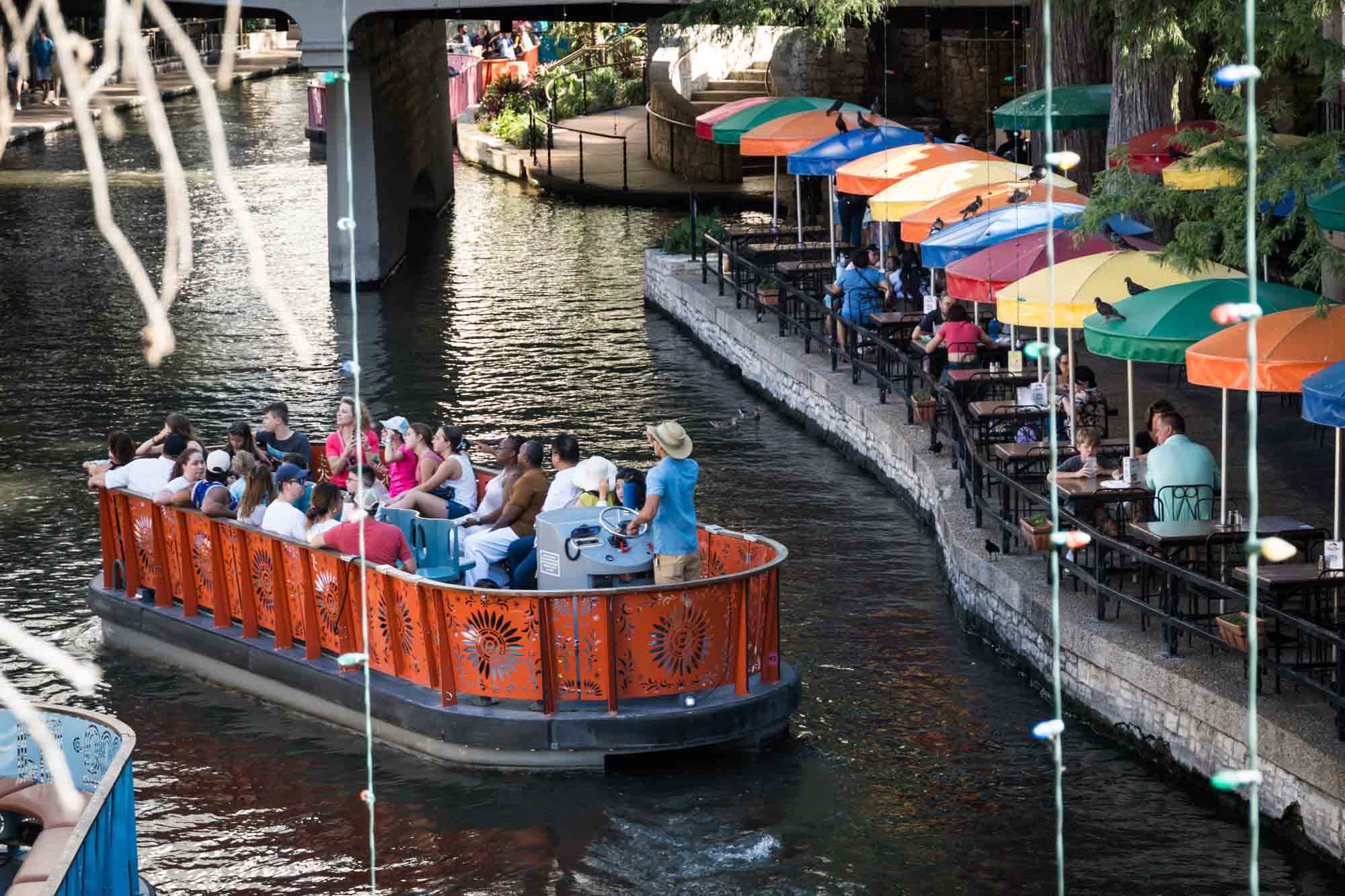 Orange boat on Riverwalk sailing past colorful umbrellas in San Antonio