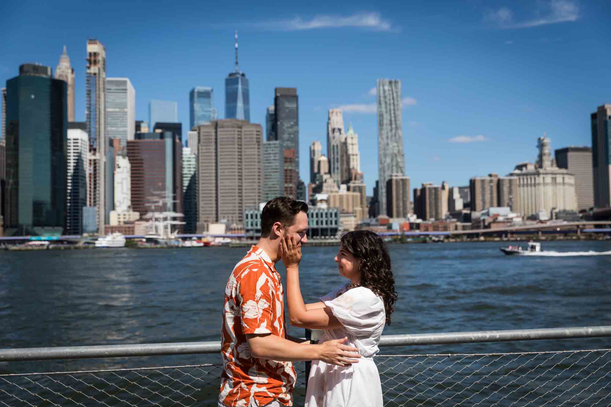 Woman touching man's cheeks in front of waterfront in Brooklyn Bridge Park