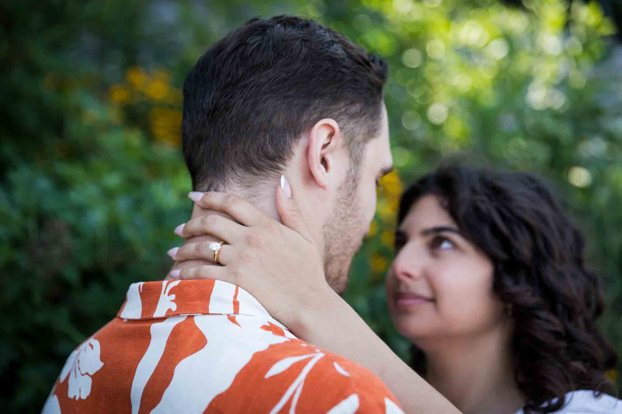Couple looking at each other with woman's hand showing engagement ring in front of green bushes in Brooklyn Bridge Park