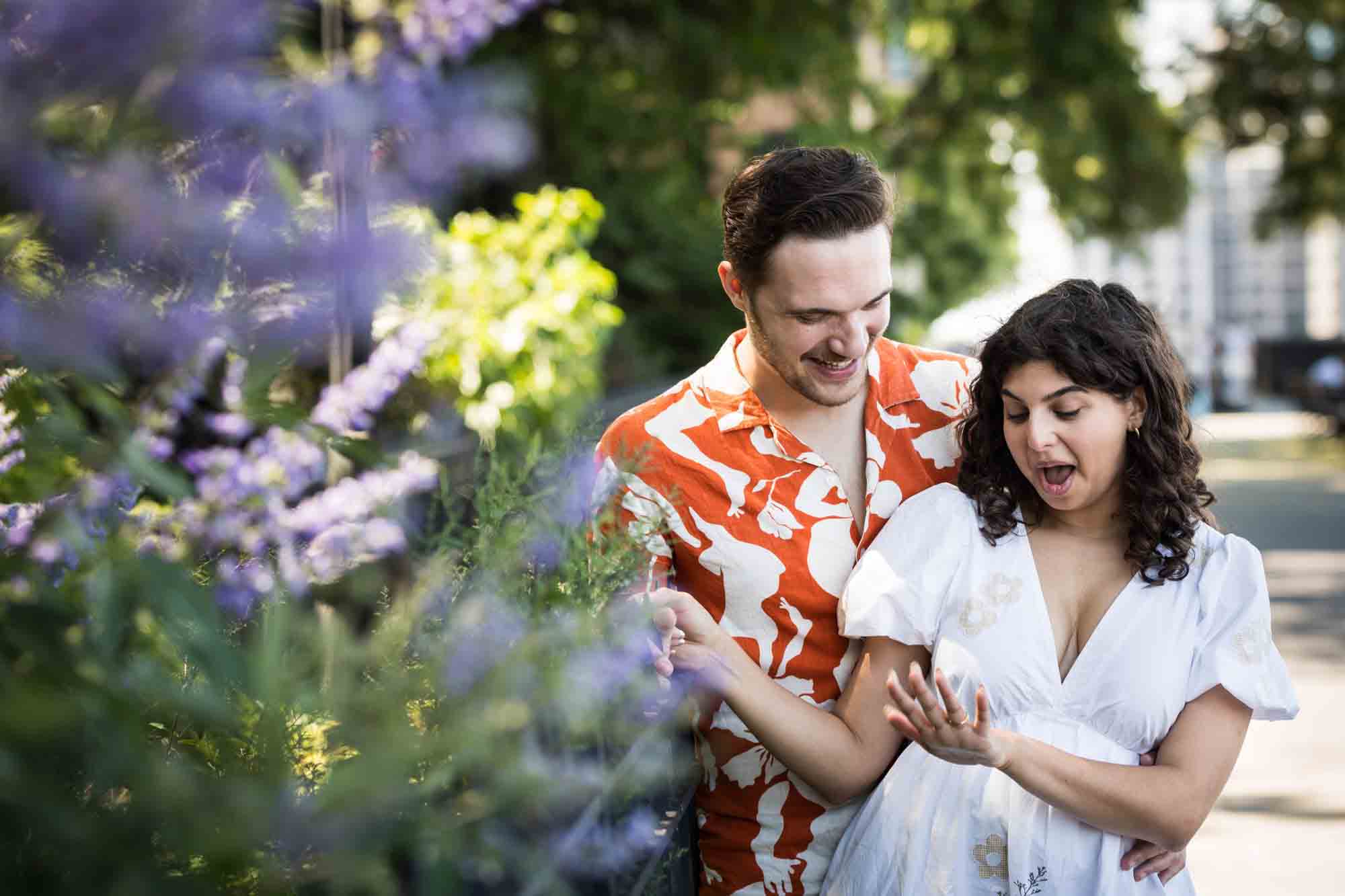 Couple looking at engagement ring on woman's finger behind purple flowers during a Brooklyn Heights surprise proposal