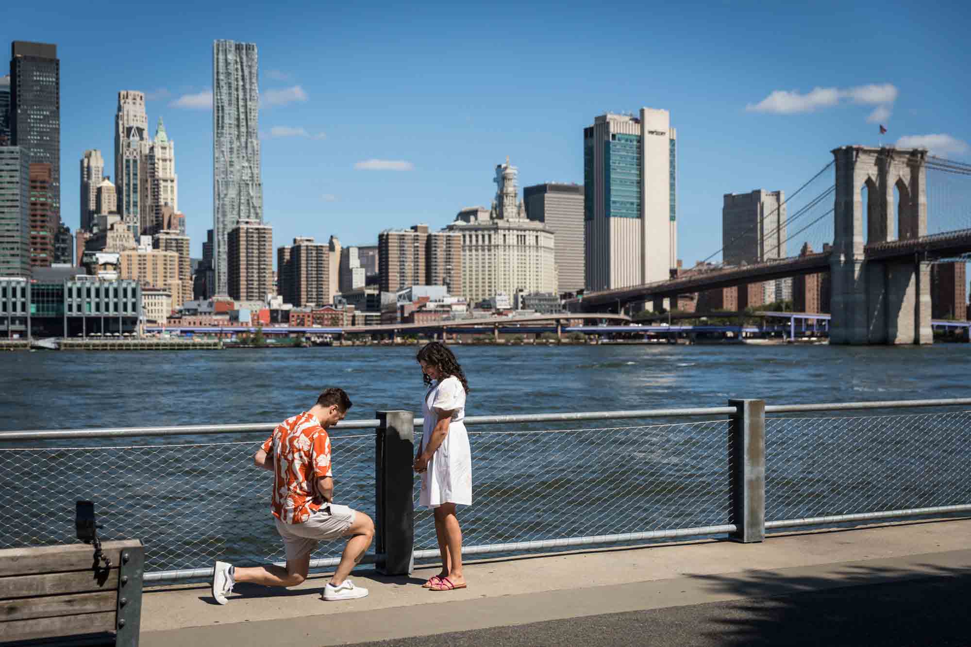 Man on one knee about to propose in front of waterfront in Brooklyn Bridge Park