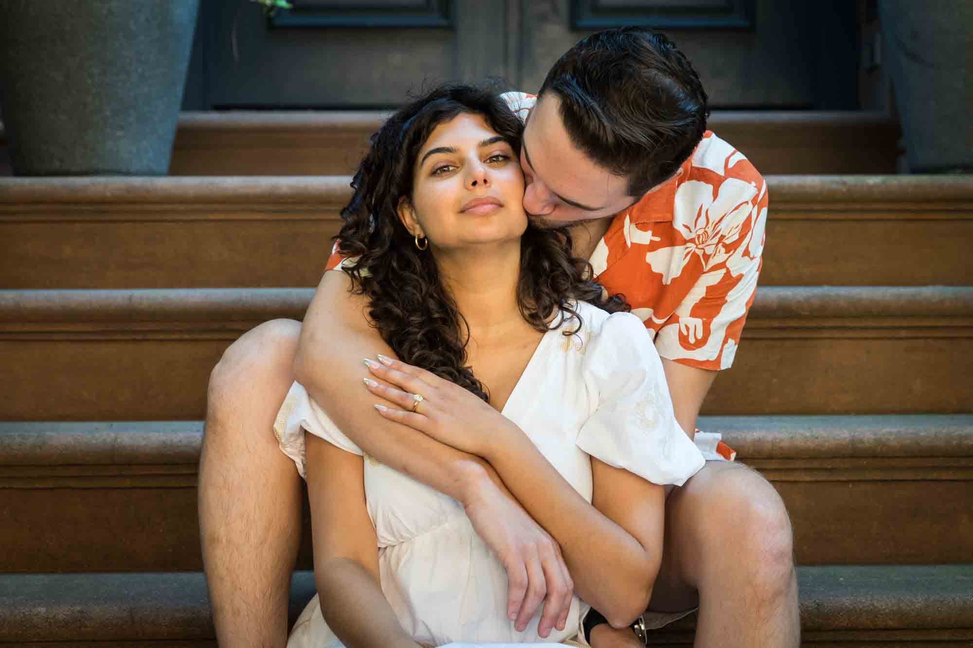 Man kissing woman on cheek on brownstone staircase during a Brooklyn Heights surprise proposal