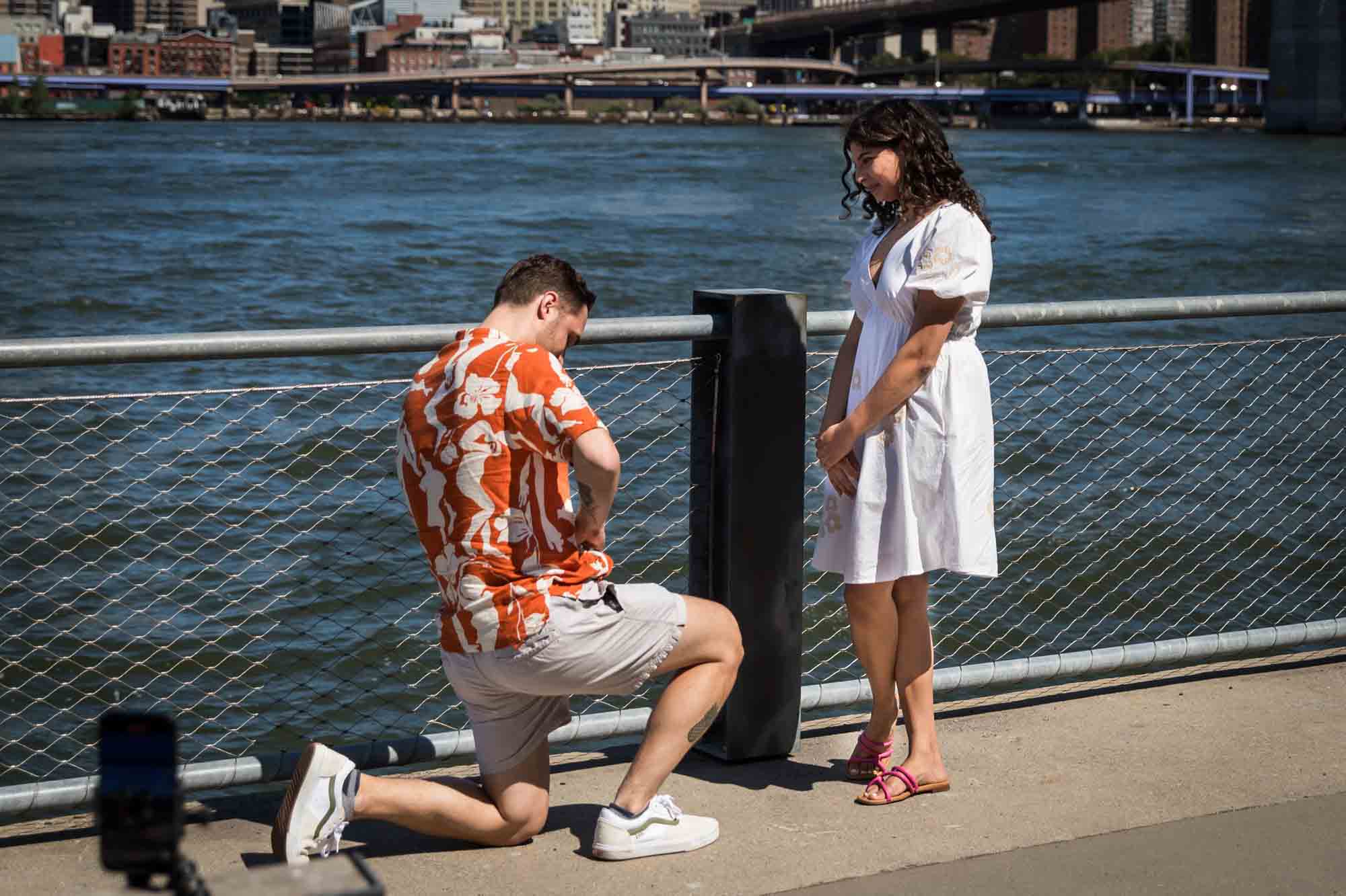 Man on one knee about to propose in front of waterfront in Brooklyn Bridge Park