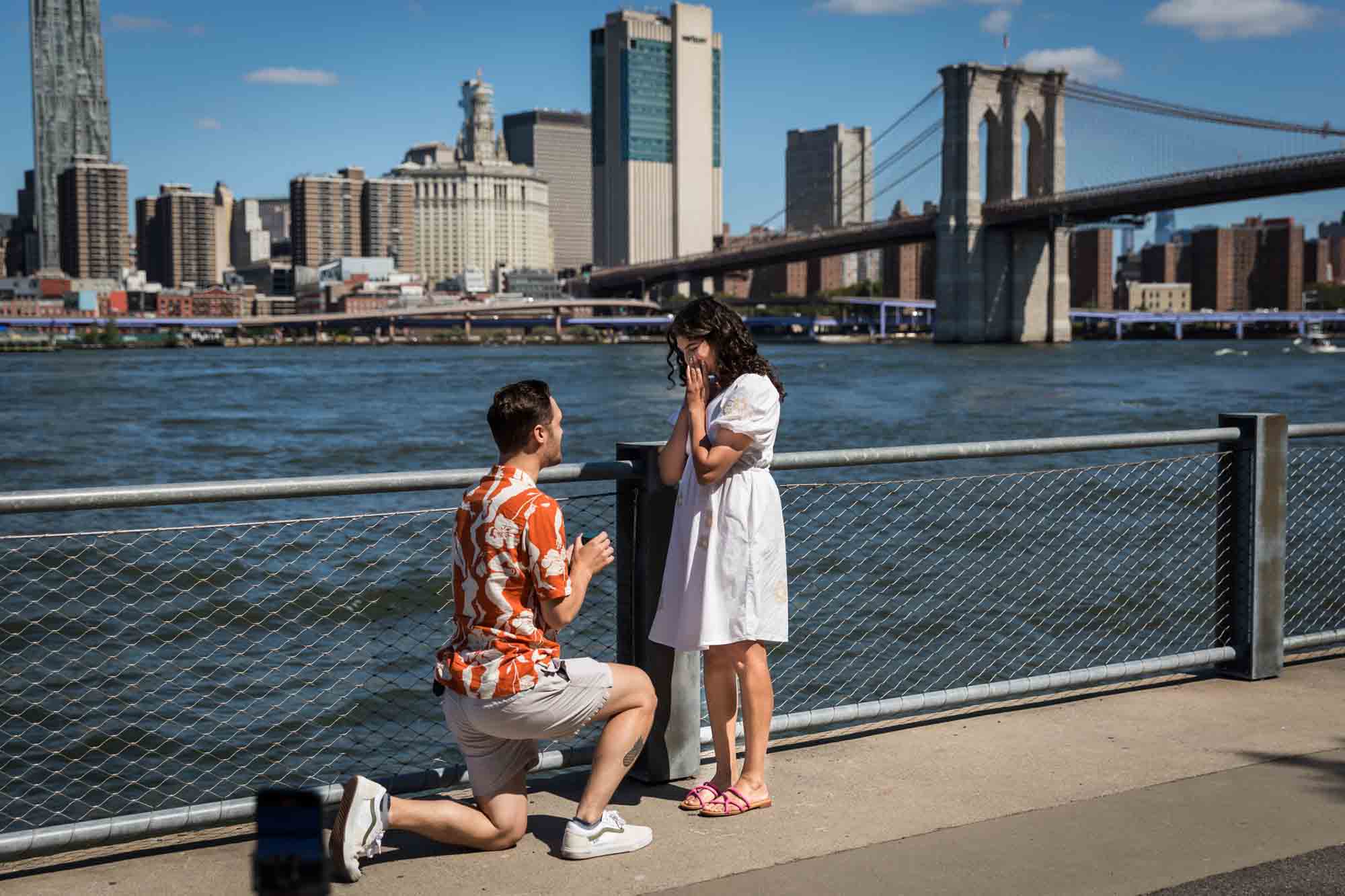 Man on one knee proposing in front of waterfront in Brooklyn Bridge Park