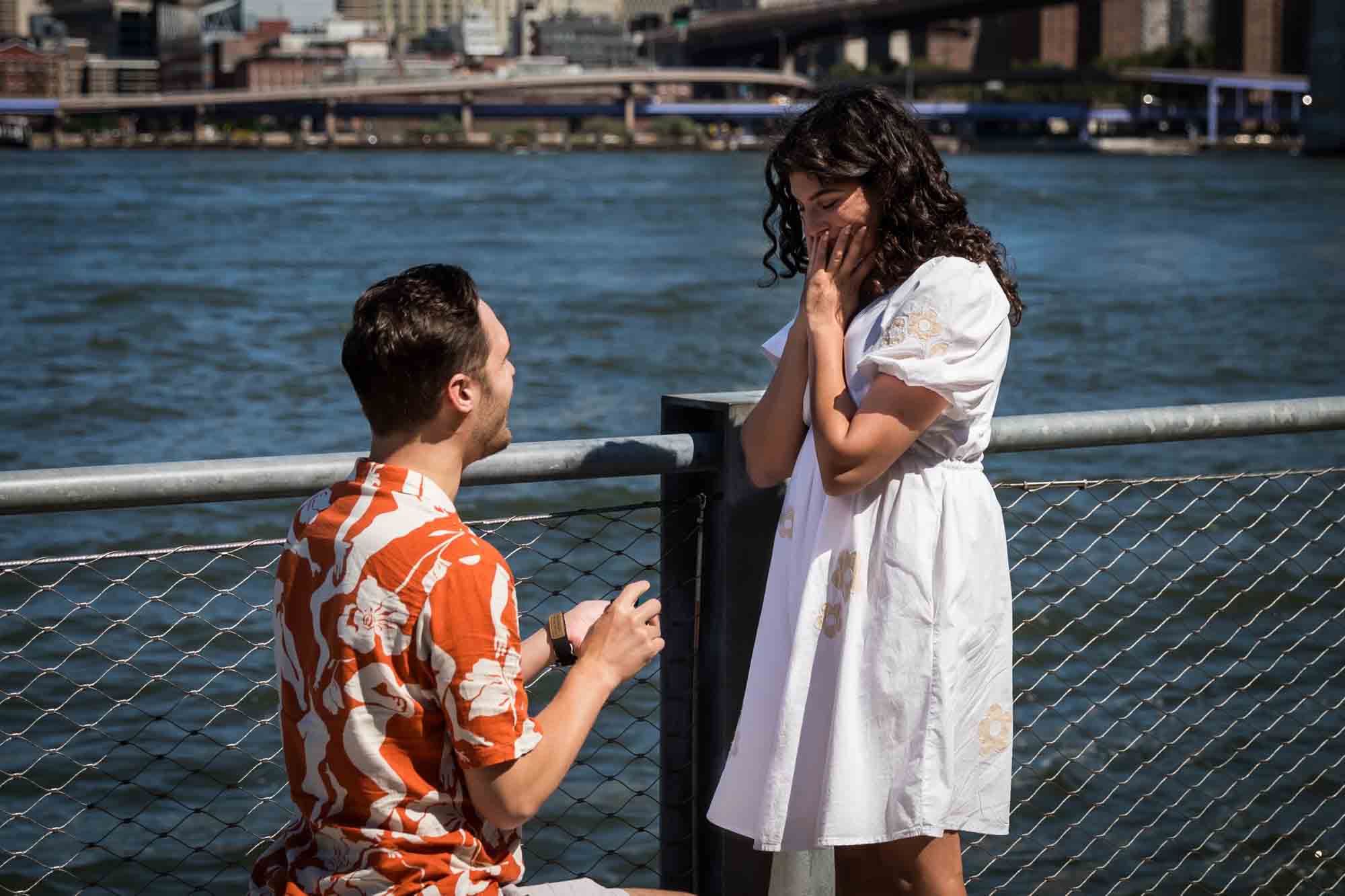Man on one knee proposing in front of waterfront in Brooklyn Bridge Park