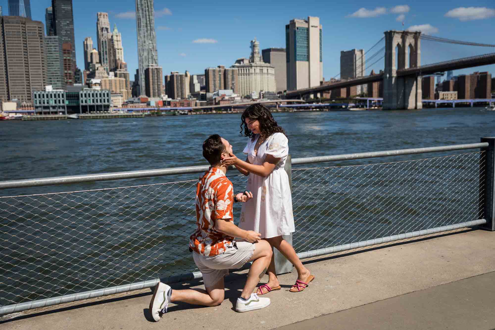 Man on one knee proposing in front of waterfront in Brooklyn Bridge Park