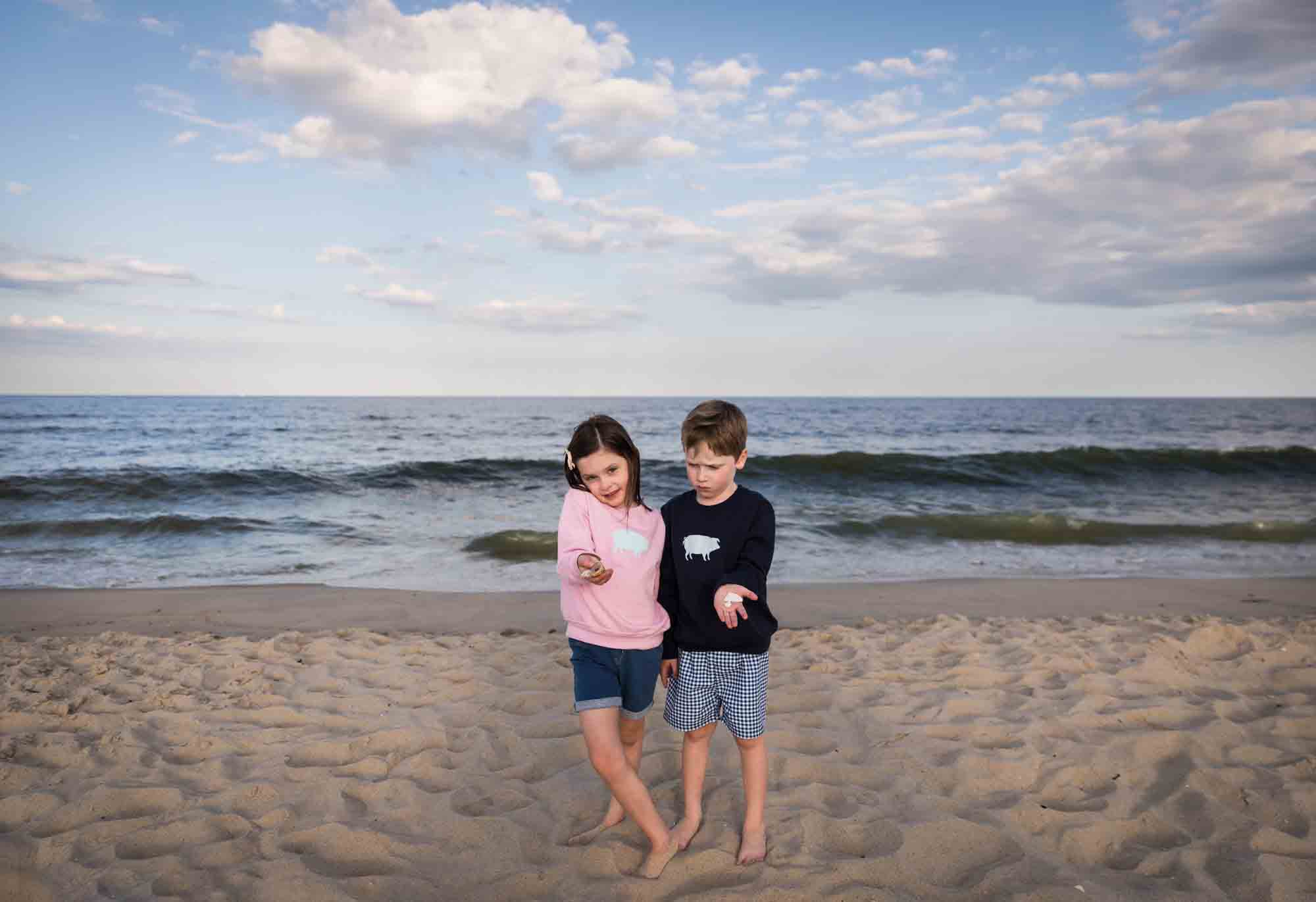 Little boy and girl with shells in hand standing on beach in front of ocean