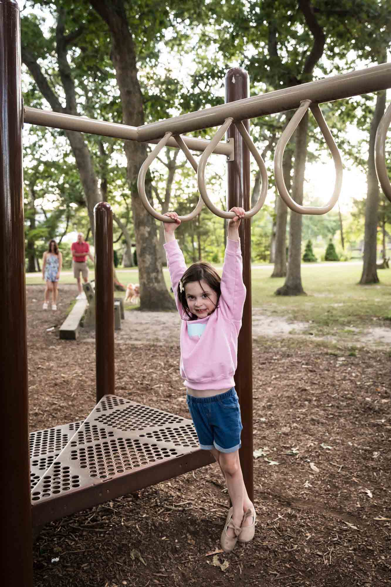 Little girl in pink sweatshirt hanging on monkey bars at playground