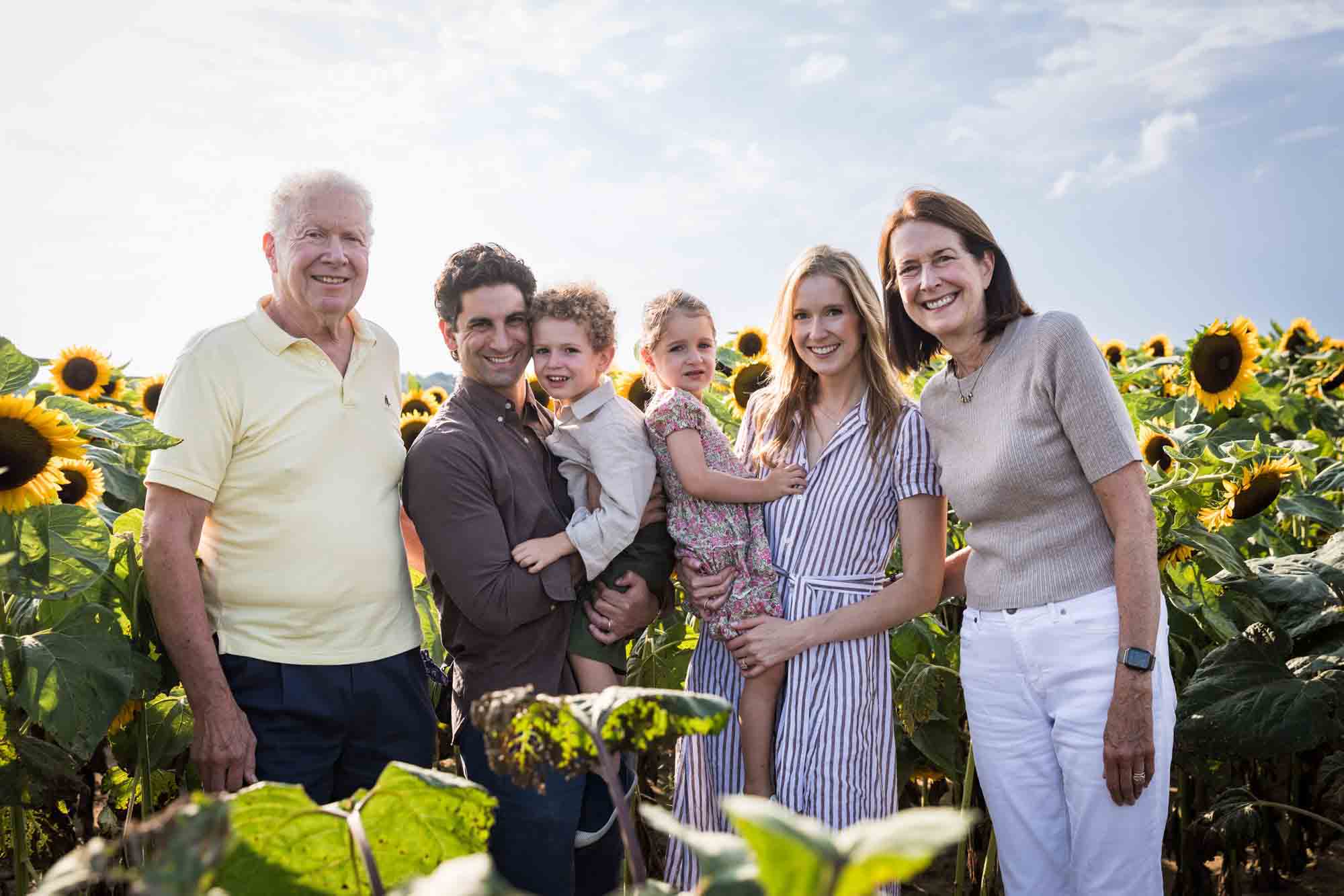 Parents holding two small children with grandparents in sunflower patch 