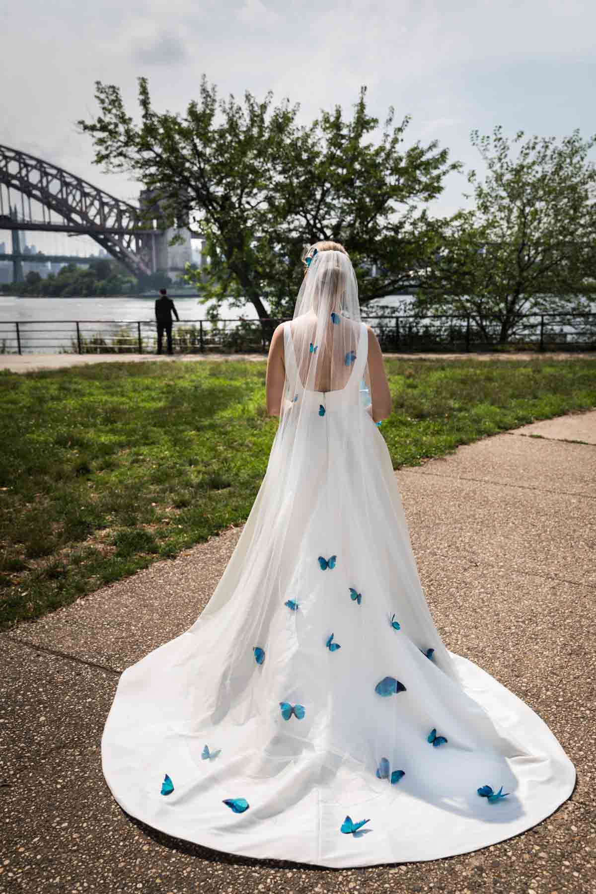 Bride standing on sidewalk looking at groom with wide train covered in butterflies in Astoria Park