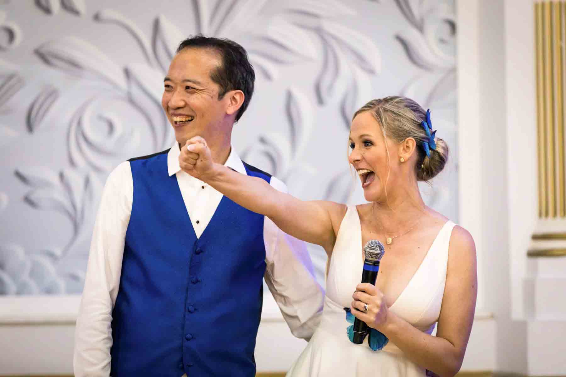 Bride holding microphone with fist raised beside groom wearing blue vest