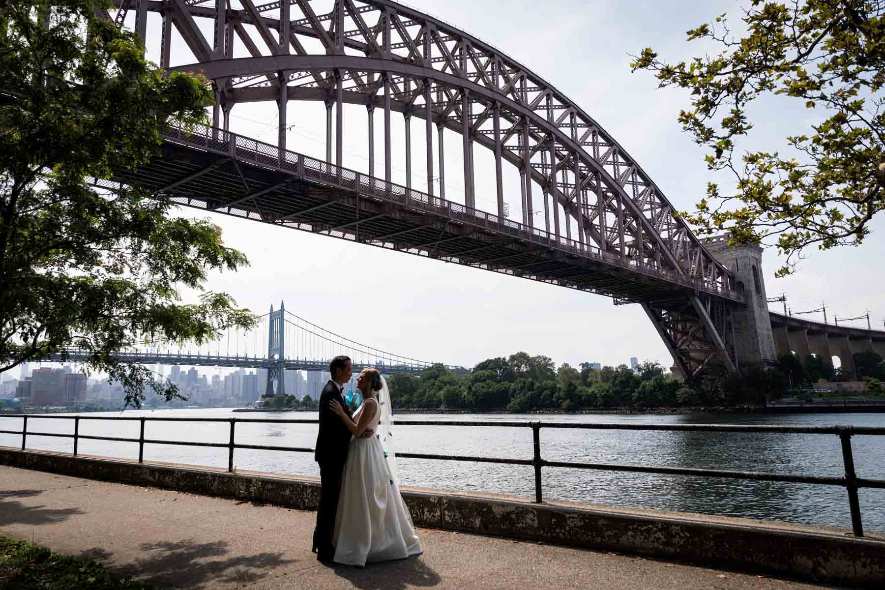 Backlit shot of bride and groom hugging along waterfront with bridges in background in Astoria Park