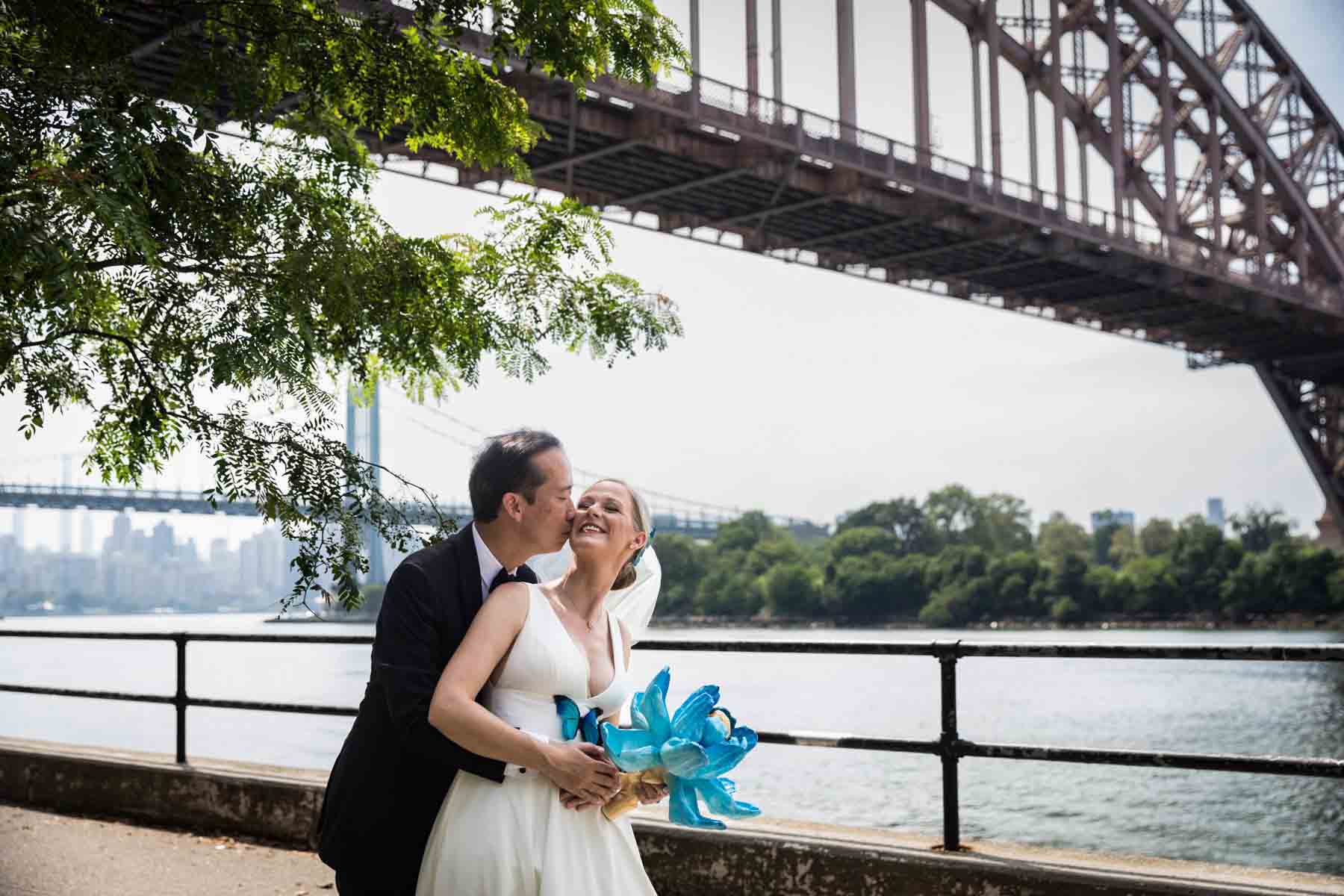 Groom kissing bride on cheek along waterfront with Hell Gate Bridge in background in Astoria Park