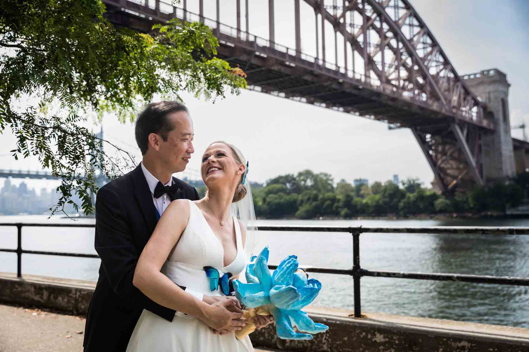Bride and groom hugging along waterfront with Hell Gate Bridge in background in Astoria Park