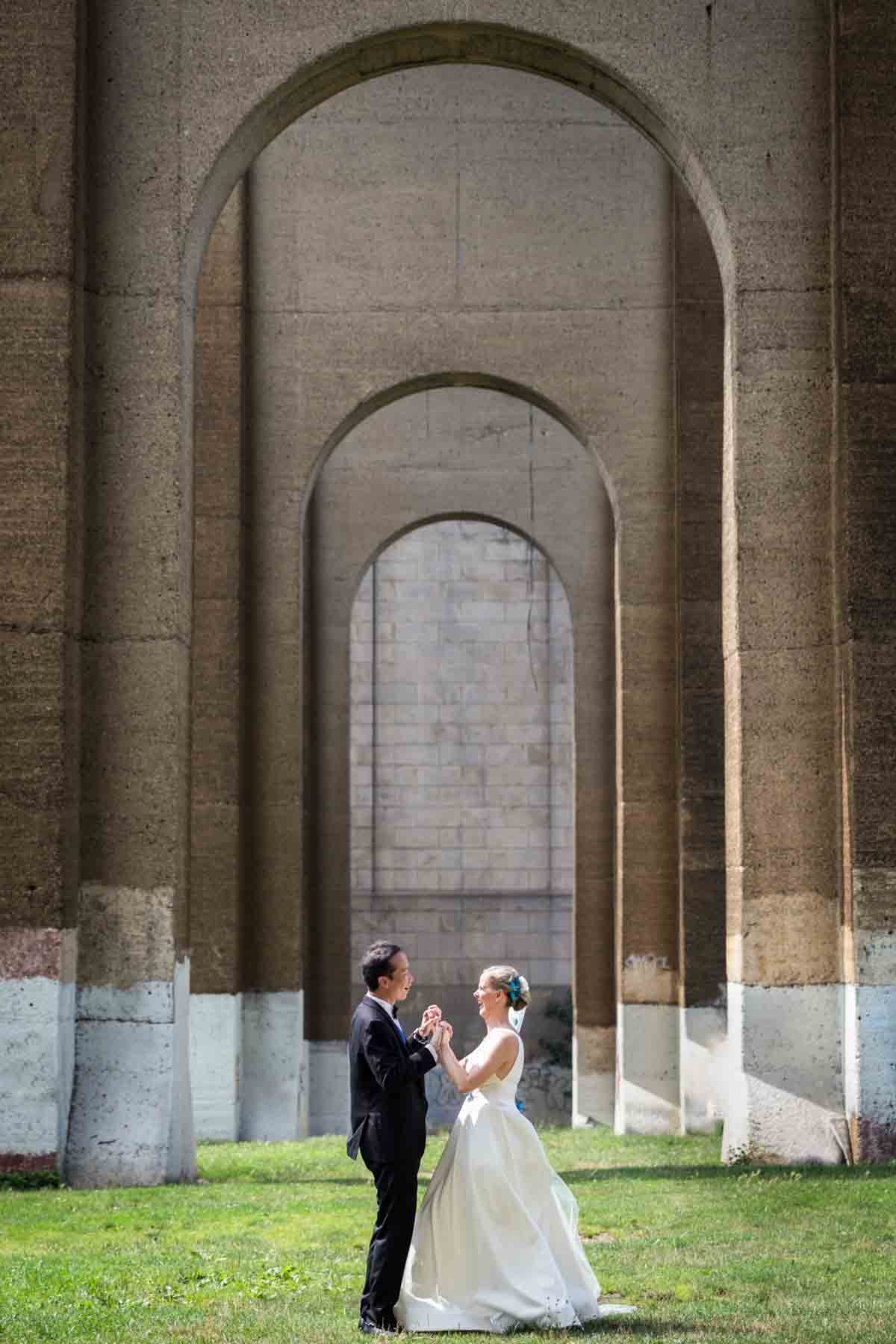 Bride and groom holding hands under Hell Gate Bridge arches in Astoria Park