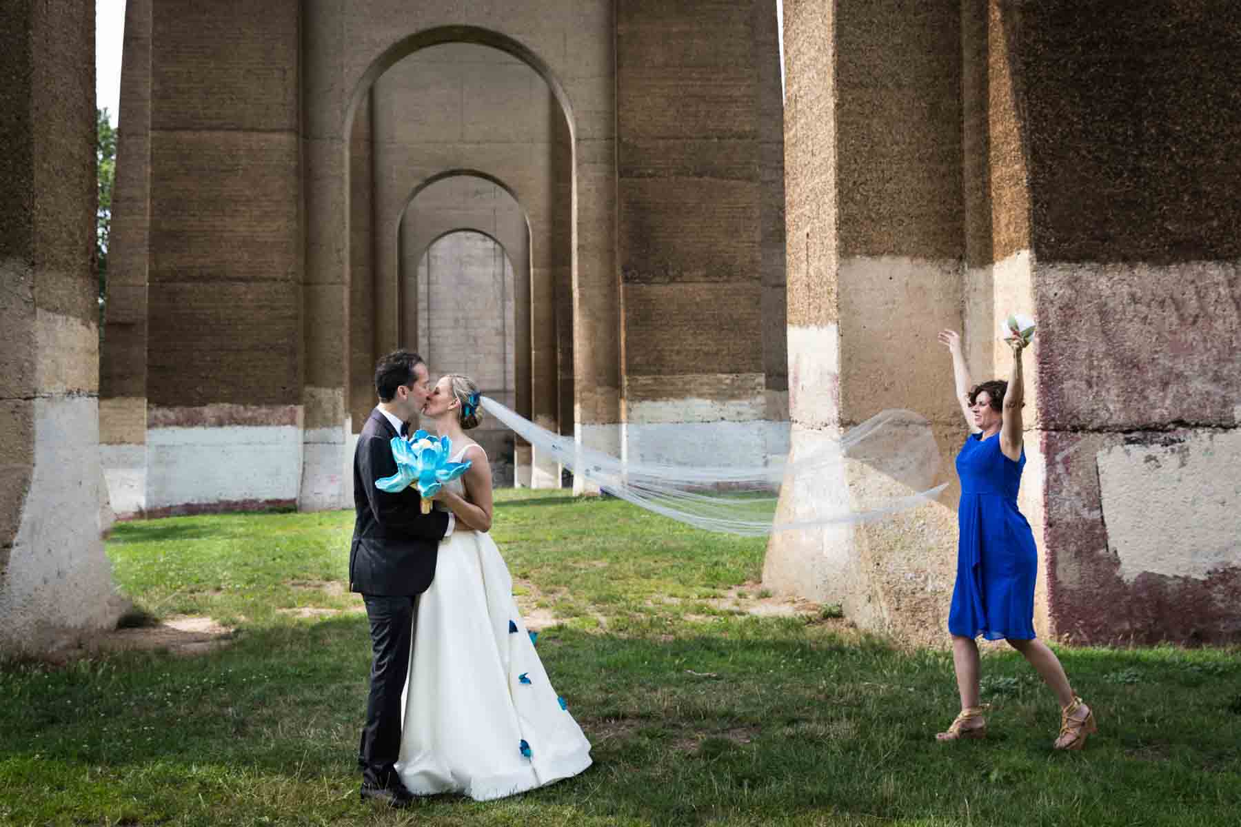 Bride and groom kissing under Hell Gate Bridge while bridesmaid in blue dress throws veil in air