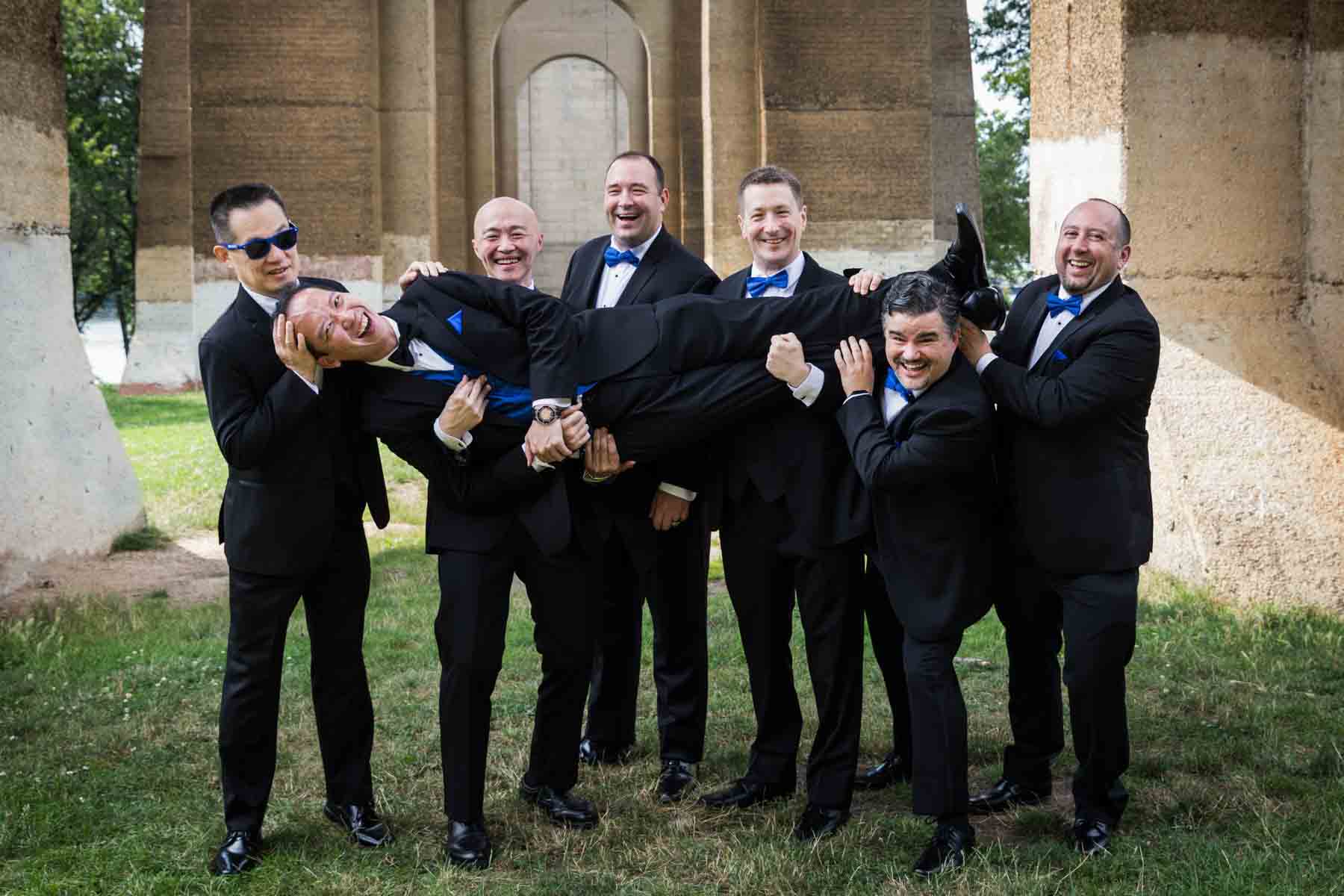 Groomsmen holding groom in the air under Hell Gate Bridge arches in Astoria Park