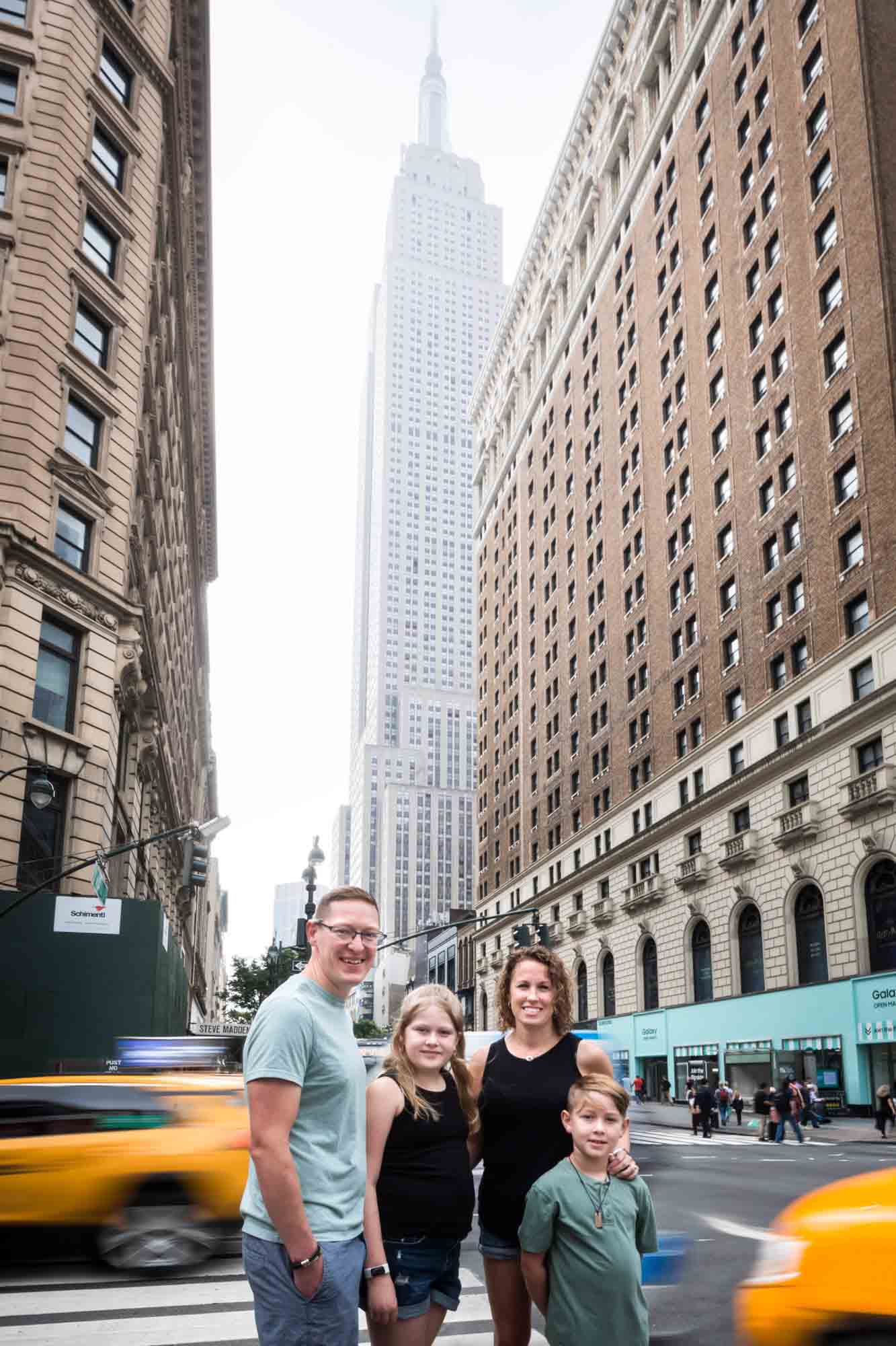 Parents and two kids standing at NYC intersection in front of speeding yellow taxis and Empire State Building 