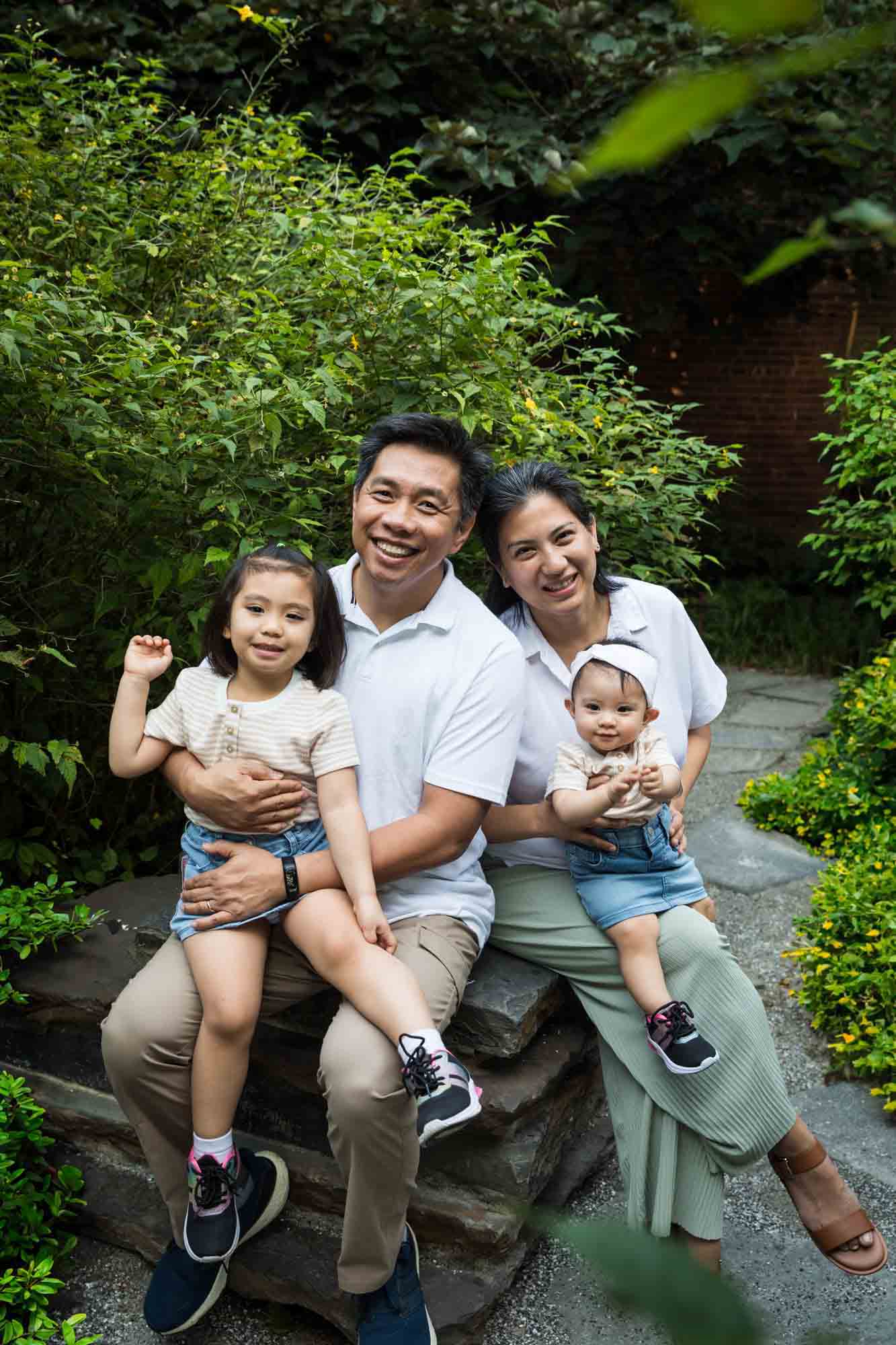 Parents sitting with two daughters on rock in front of bushes for an article on Brooklyn Bridge Park family portrait tips for young children