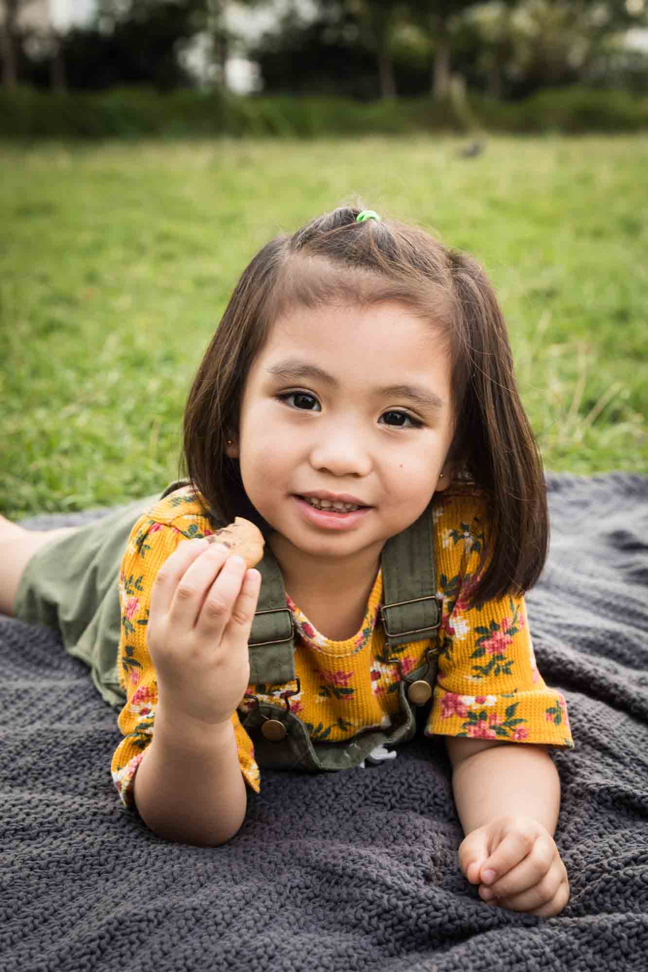 Little girl wearing yellow shirt eating cookie on grey blanket