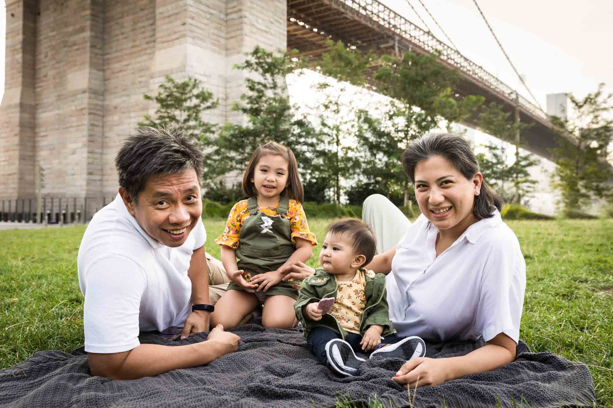 Parents with two young daughters on grey blanket for an article on Brooklyn Bridge Park family portrait tips for young children