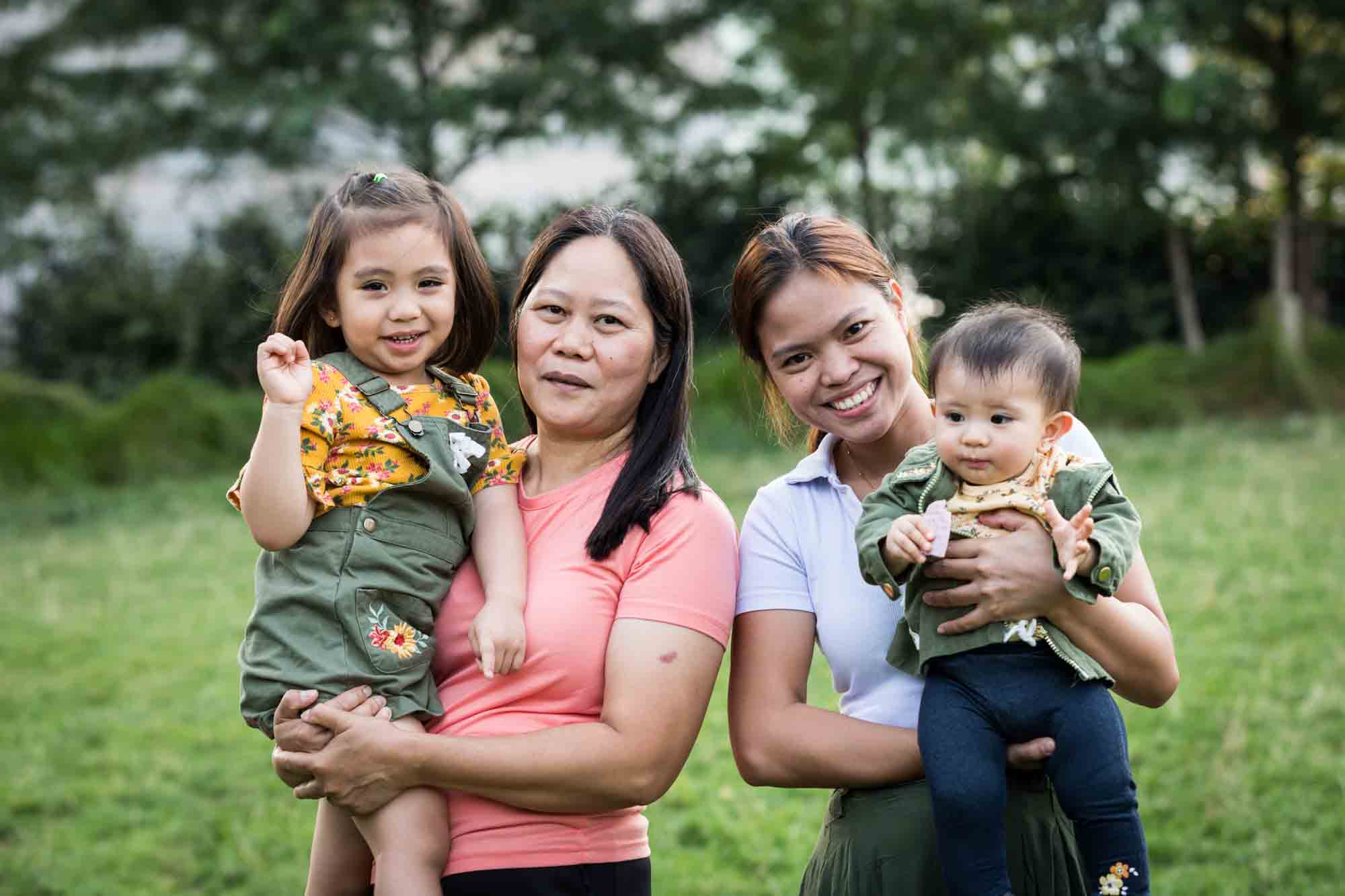 Two women holding little girls in front of grass and trees in Brooklyn Bridge Park